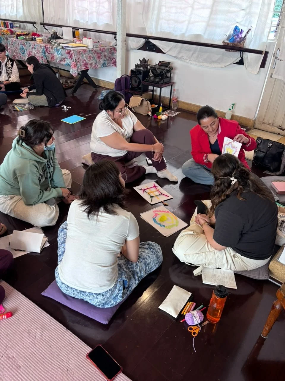 Grupo de personas sentadas en círculo en el suelo participando en una sesión creativa o taller, con materiales de arte y libros alrededor en un espacio interior