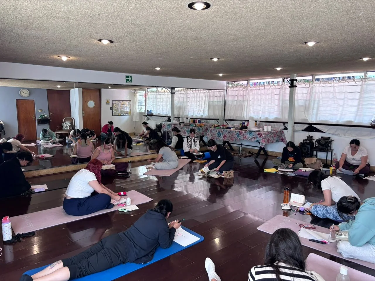 Grupo de personas practicando arteterapia en interior, sentadas en colchonetas, en un espacio con madera en el suelo y cortinas en las ventanas.