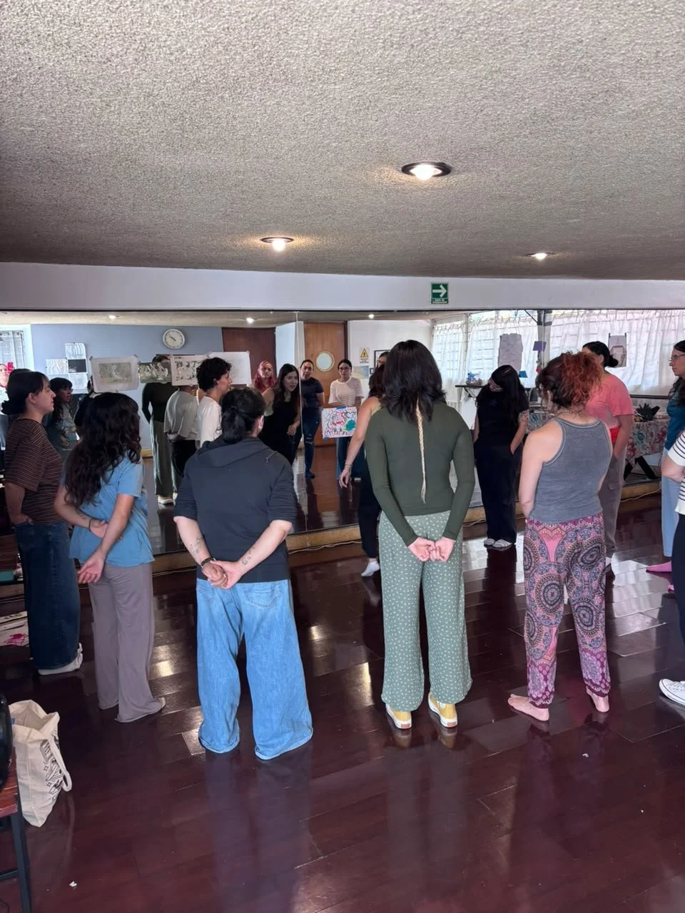 Grupo de personas en una clase o taller, de espaldas, algunas con brazos cruzados, observando a una instructora frente a un espejo, en un ambiente interior con suelo de madera y luz artificial.