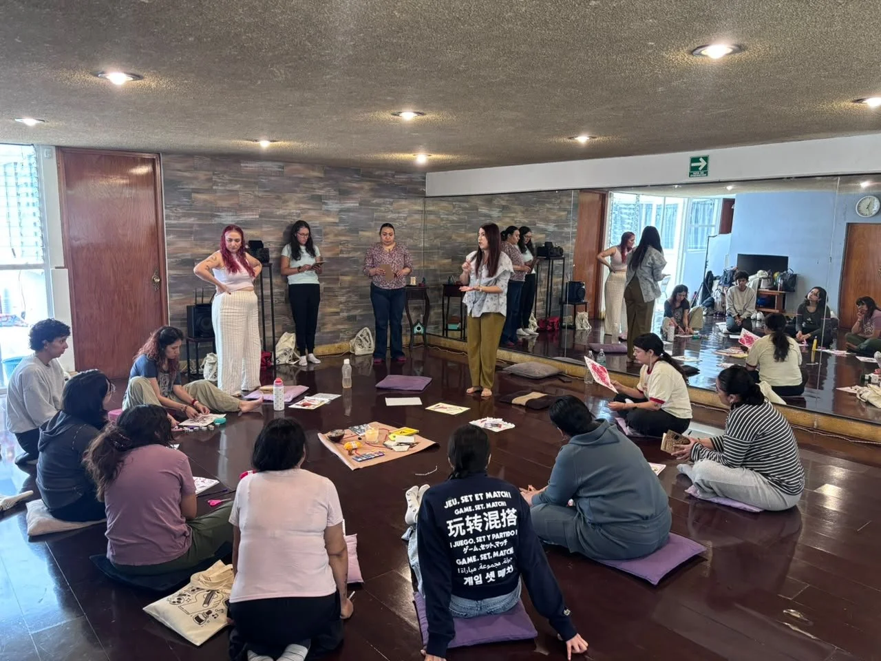 Grupo de personas sentadas en el suelo en un taller, algunas tomando notas, y varias mujeres de pie en un escenario, una de ellas hablando.