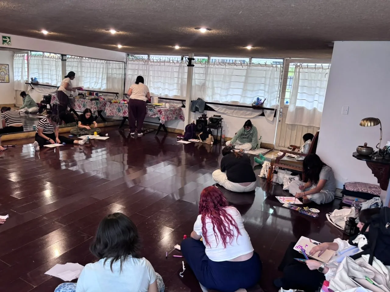 Varias personas sentadas en el suelo, participando en un taller de arte o manualidades en una habitación con suelo de madera y cortinas blancas.