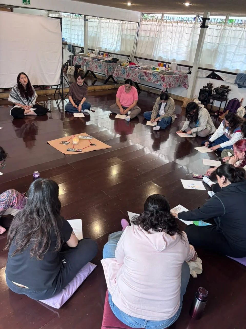 Un grupo de personas sentado en círculo en un espacio interior con piso de madera, participando en una actividad grupal. Algunos tienen libros en las manos, y hay mesas con productos y materiales en el fondo.