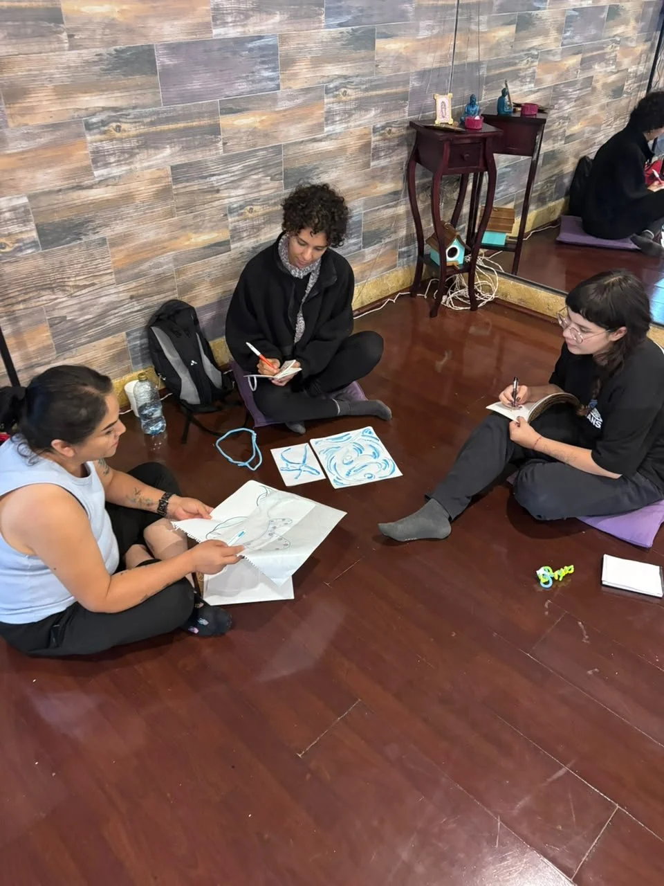 Tres personas sentadas en el suelo discutiendo y dibujando, con obras de arte y objetos personales alrededor.