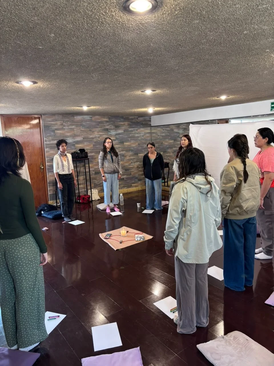 Grupo de personas de pie en círculo en una sala, participando en una actividad o taller, con objetos y notas en el suelo.