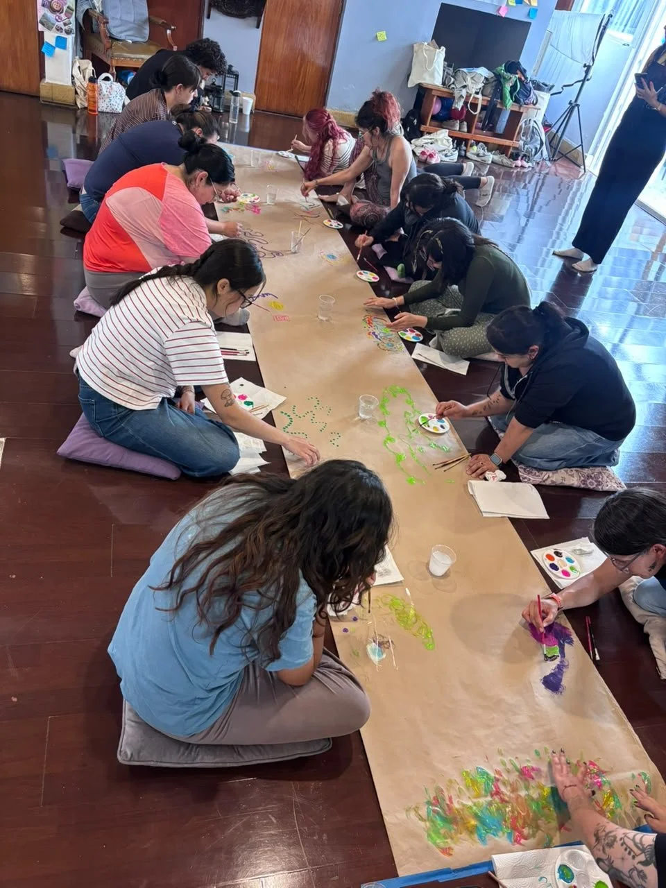 Varias personas sentadas en el suelo pintando en una gran hoja de papel con acuarelas y pinceles, en un ambiente interior.