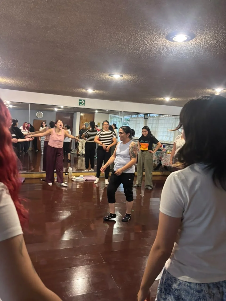 Grupo de mujeres bailando y riendo en un estudio con pisos de madera y espejo grande, parece ser una clase o reunión social.
