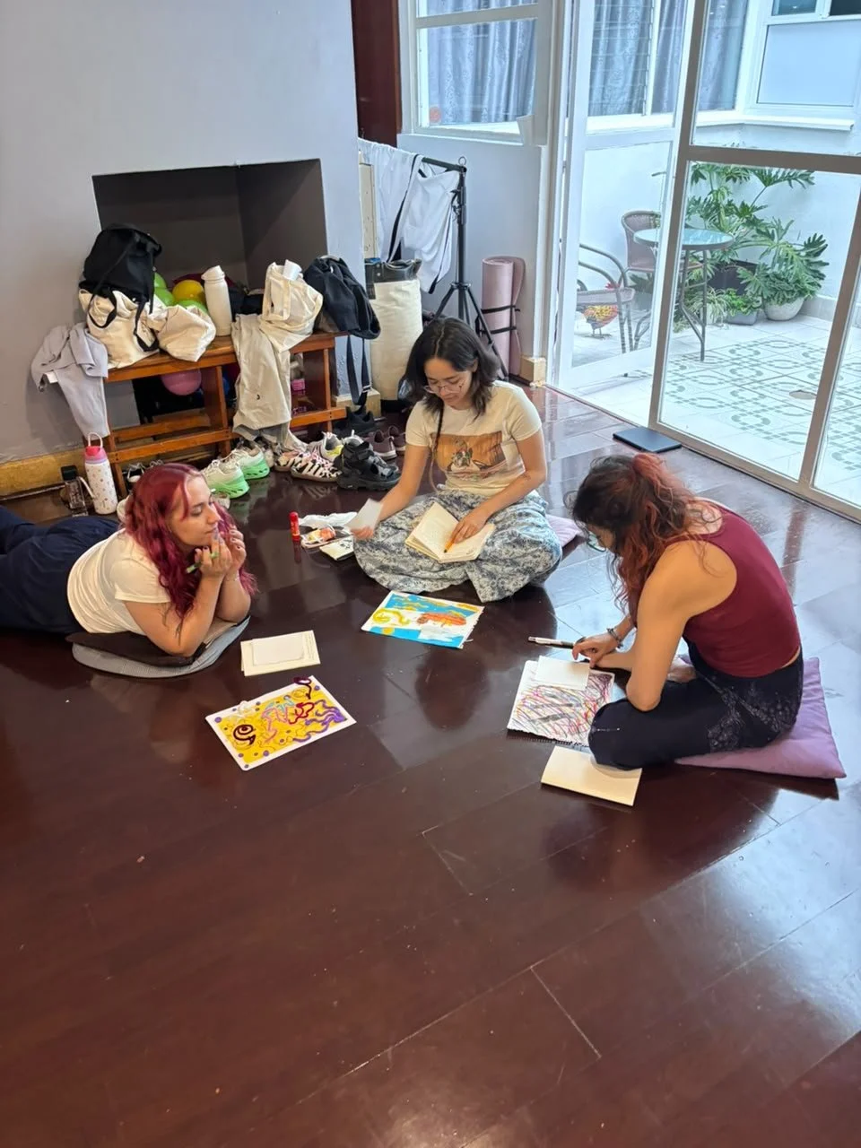 Tres personas sentadas en el piso de una habitación, rodeadas de libros, dibujos, y artículos de arte, participando en una actividad creativa o de estudio.