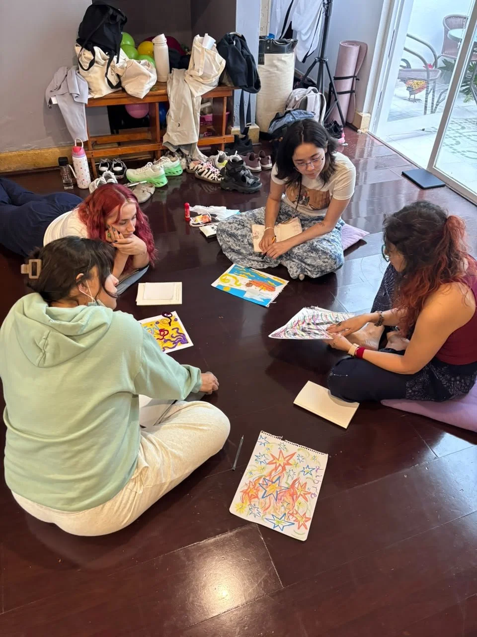 Cuatro personas sentadas en el suelo, leyendo y discutiendo dibujos y libros en una habitación. Hay ropa y calzado en una mesa y en el suelo, así como bolsas y objetos personales en el fondo.