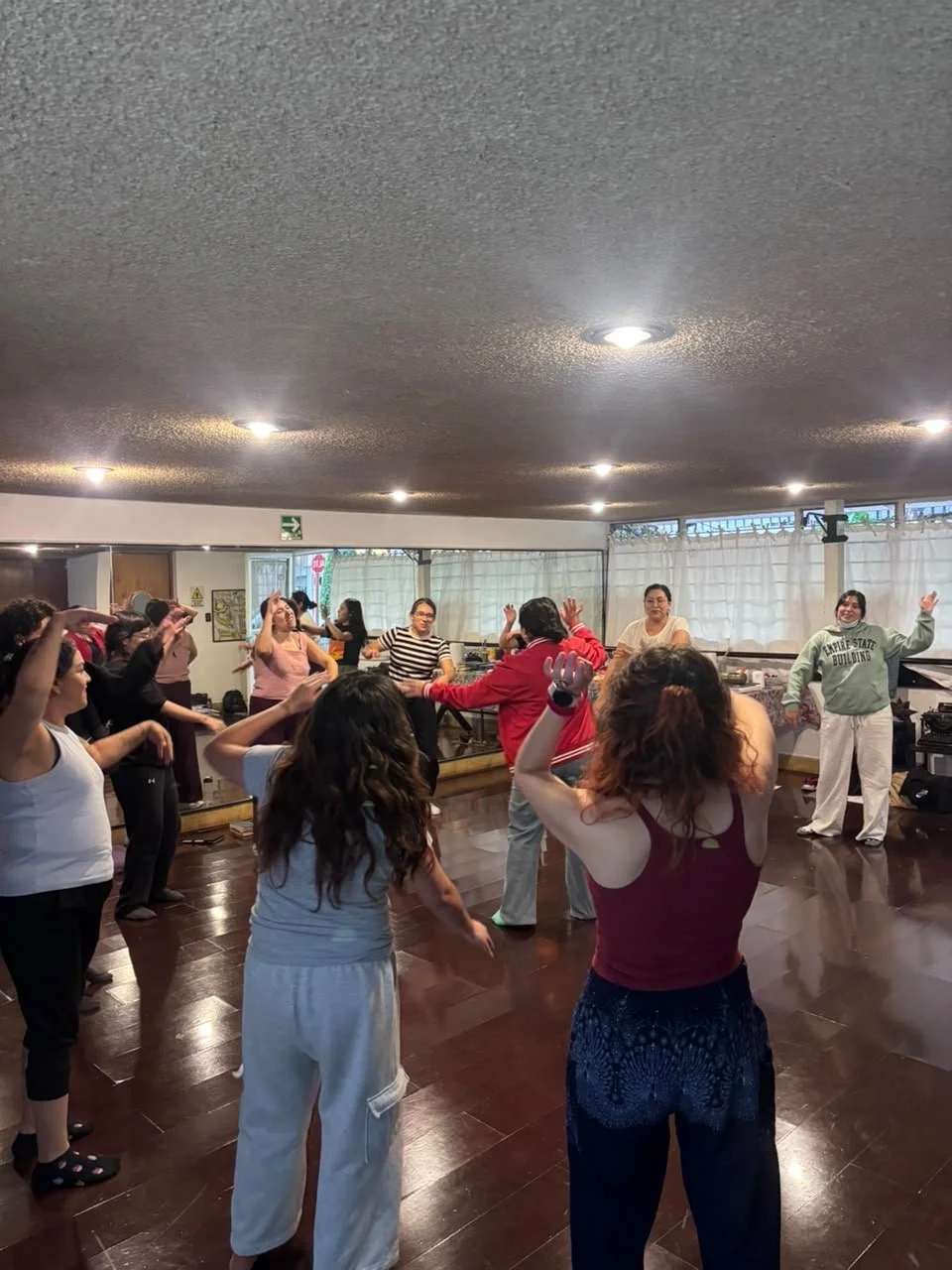 Grupo de personas bailando en una sala con piso de madera y cortinas blancas en las ventanas.