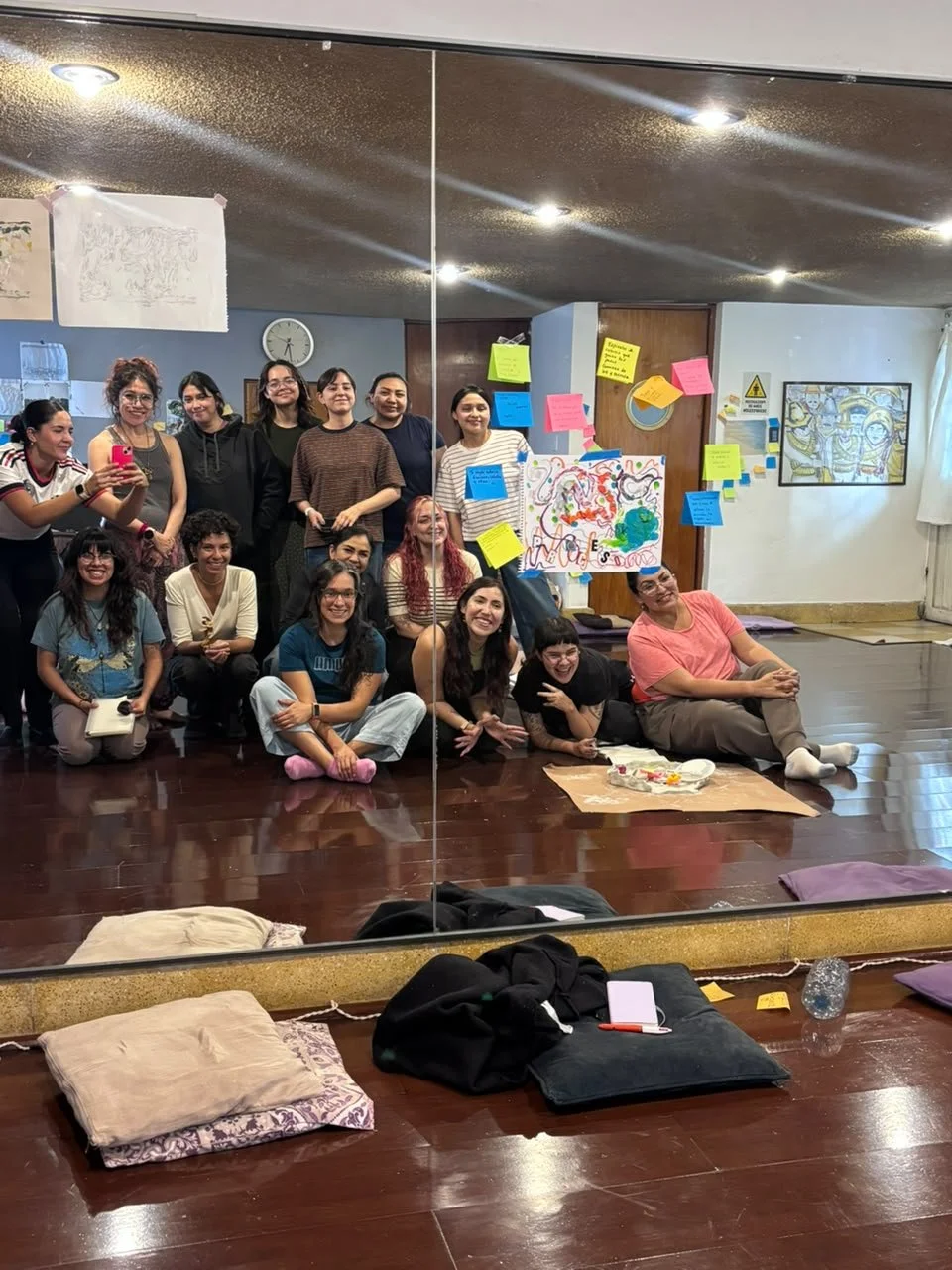 Grupo de personas en una sala de clase, algunas sonrientes y otras tomando una selfie con un espejo, con notas adhesivas de colores en la pared y un dibujo en cartulina. En el suelo hay cojines, una botella de agua y objetos personales.