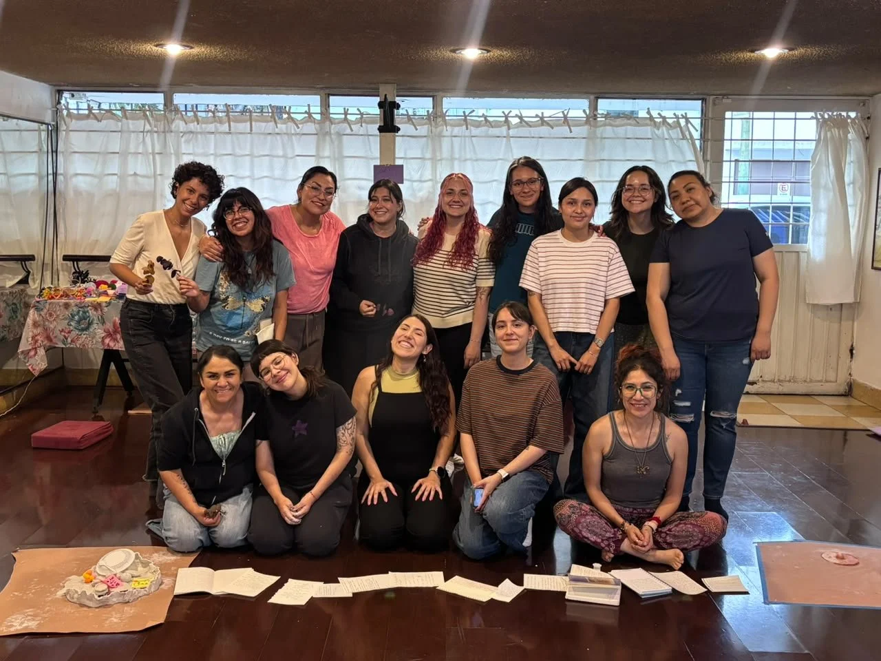 Grupo de mujeres sonriendo en un ambiente interior, algunas están sentadas y otras de pie, con mesas y objetos decorativos en el fondo.