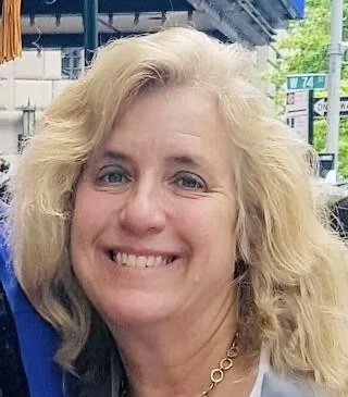 Close-up of a smiling blonde woman with curly hair, standing outdoors in an urban environment.