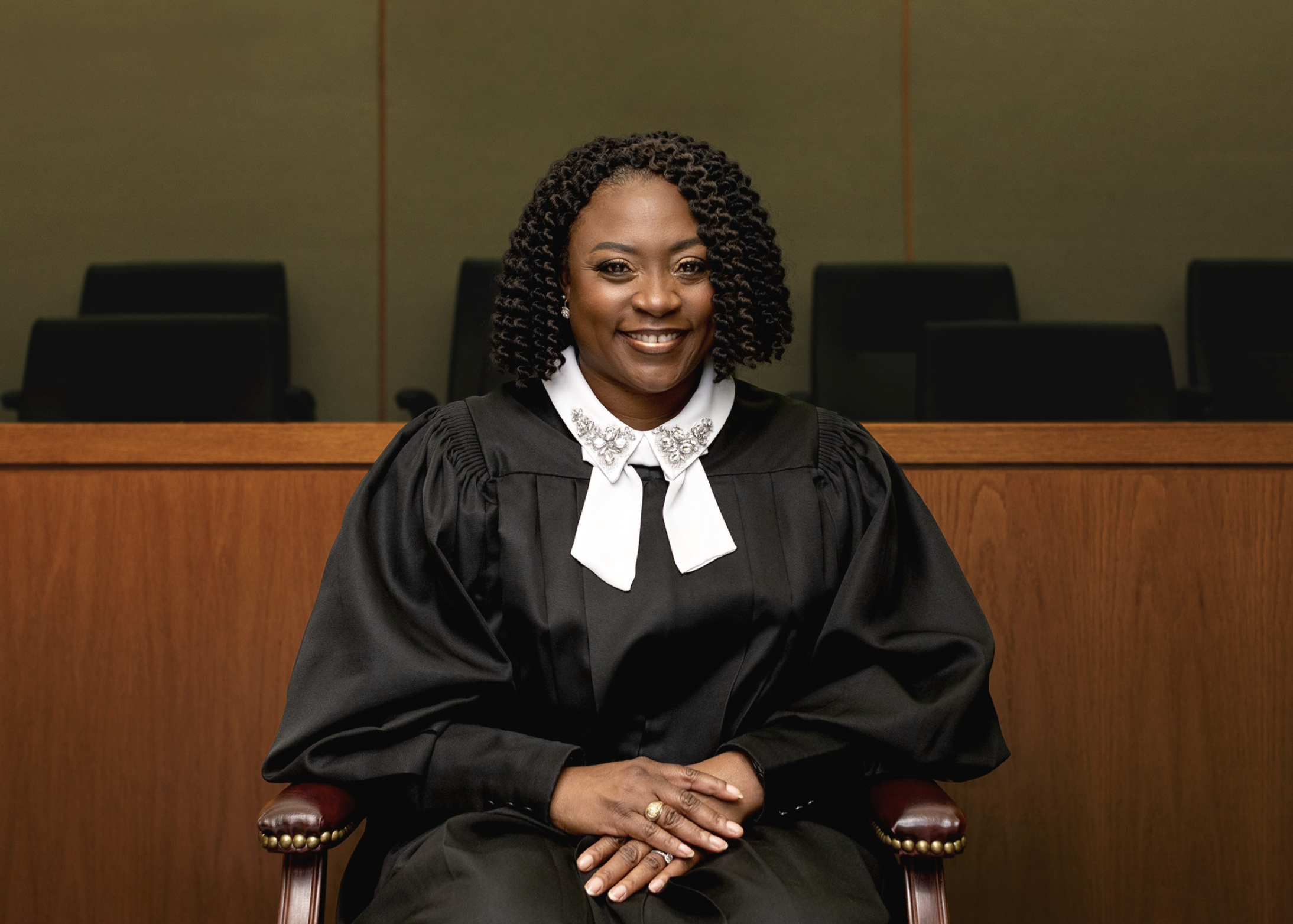 A woman with curly hair wearing a judge's robe and sitting on a wooden chair in a courtroom.
