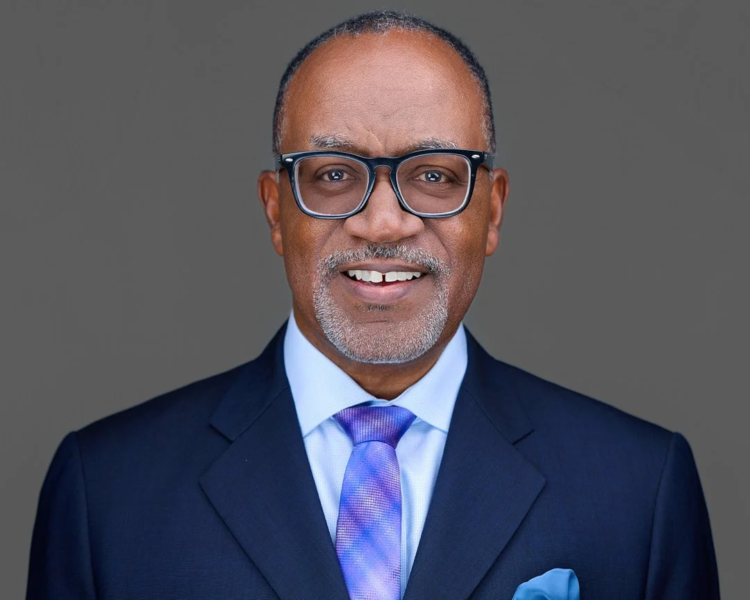 An African-American man with glasses, a suit, and a tie, smiling against a gray background.