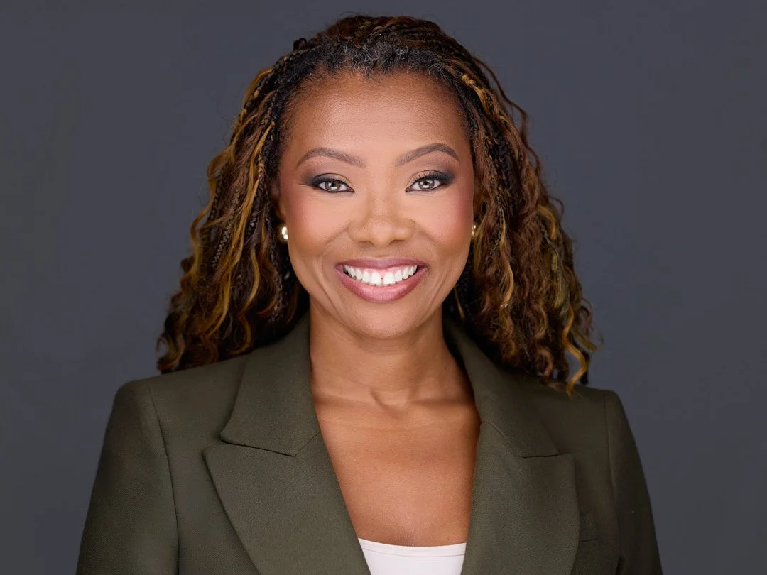 A professional woman with curly hair, smiling, dressed in a dark blazer and white top.