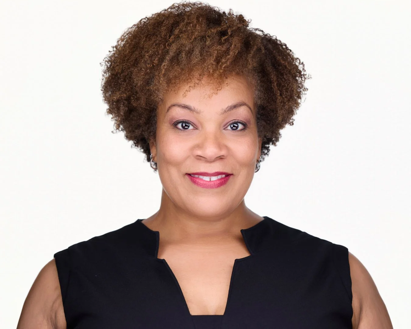 A woman with curly brown hair, wearing a black sleeveless top, smiling against a white background.