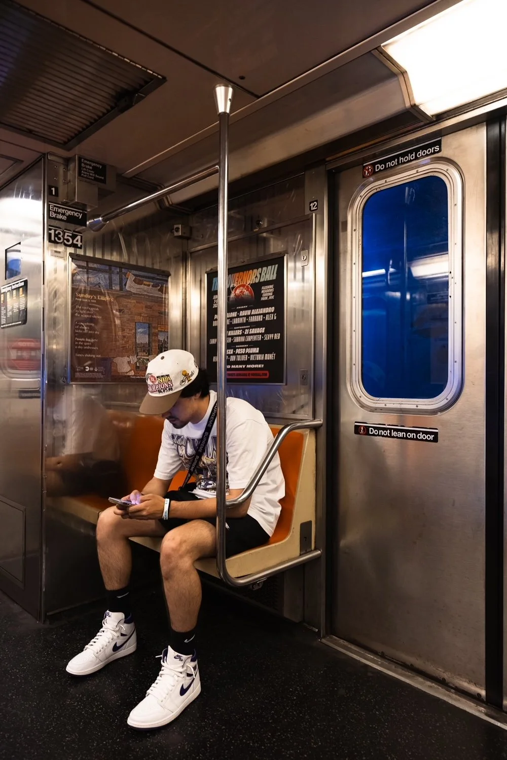 A young man sitting on a subway train seat, looking at his phone. He is wearing a white cap with sports logos, white sneakers, black shorts, and a white T-shirt. The subway interior has orange seats, a pole in front of him, and a blue window on the door with multiple signs and posters around.