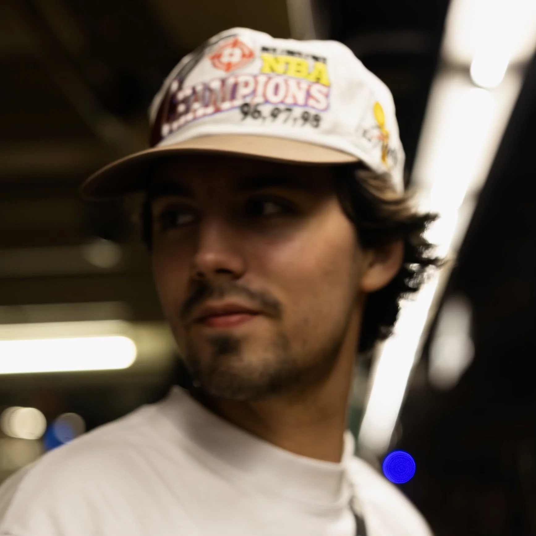 Partial view of a person wearing a hat with various patches and emblems, including one that reads "CHAMPIONS 96-97-98" and another that reads "NBA". The person is looking to the side with a neutral expression, in an indoor setting with blurred lights in the background.