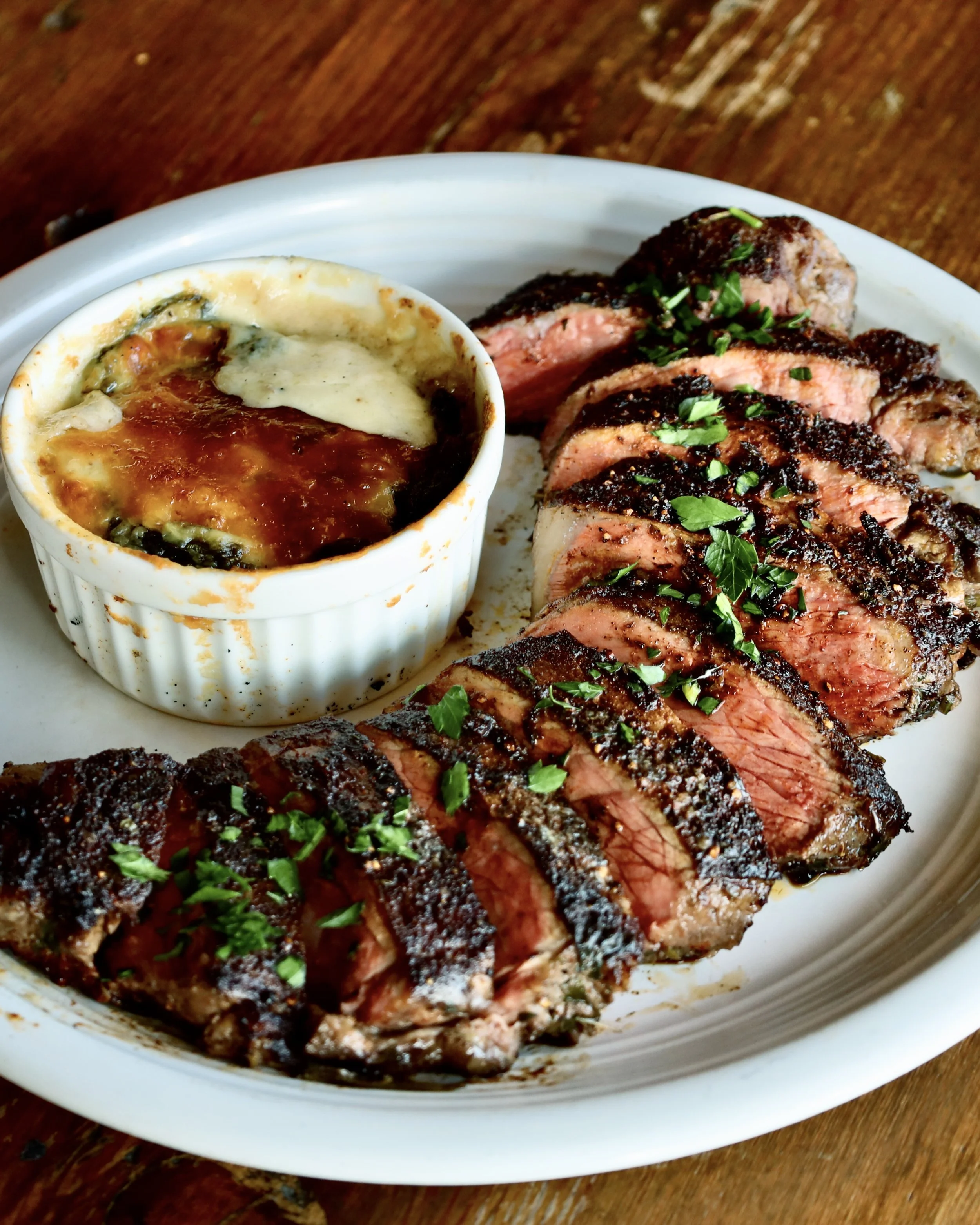 Blackened NY Strip Steak seared and sliced alongside a white ramekin with cheesy spinach scalloped potatoes.