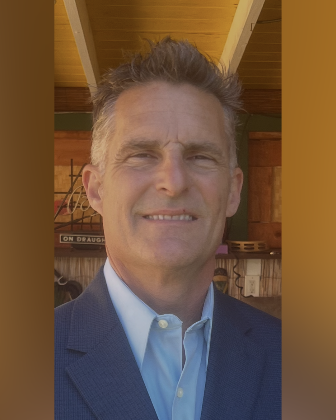 A middle-aged man with short, gray hair, dressed in a blue suit and white shirt, smiling in a restaurant or café with wooden walls and ceiling.
