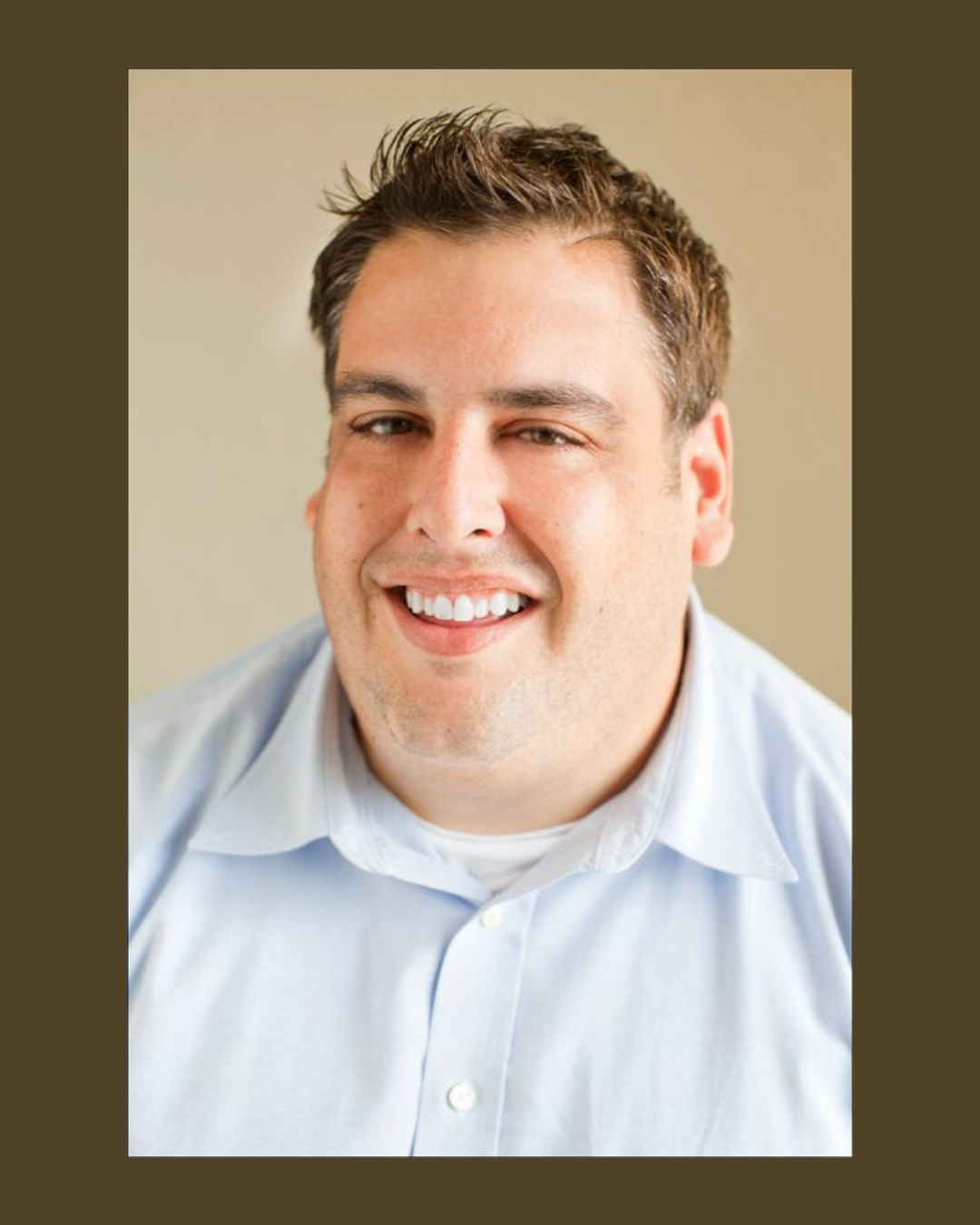 Close-up of a smiling man with short dark hair, wearing a light blue dress shirt, against a neutral background.