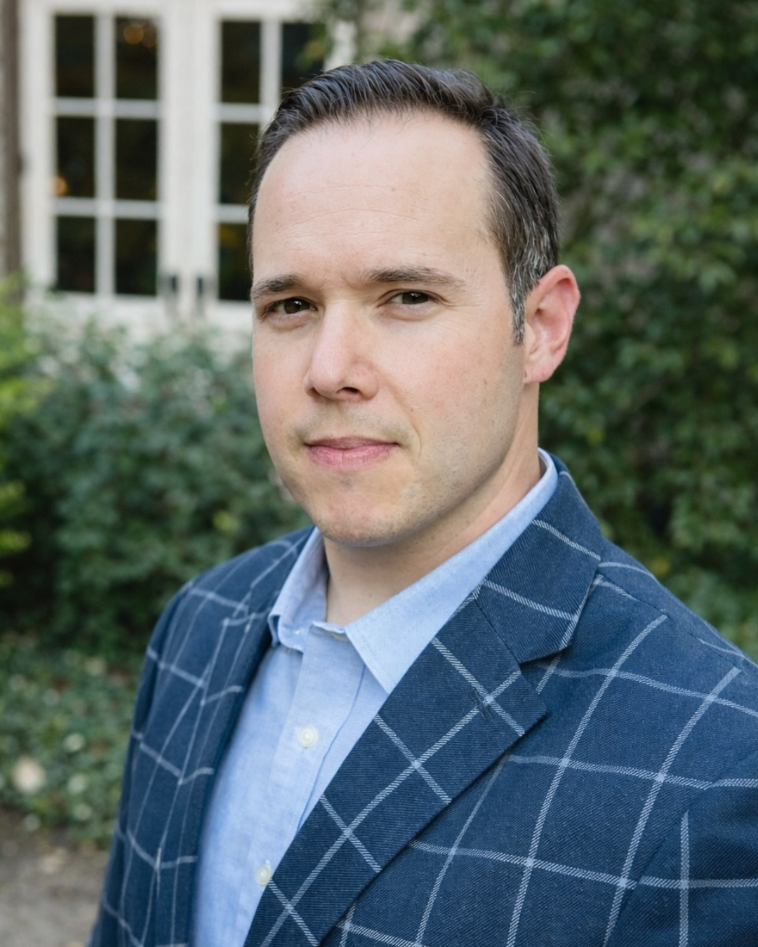 Portrait of a man with short dark hair, wearing a blue checkered blazer and light blue shirt, standing outdoors with greenery and a house in the background.