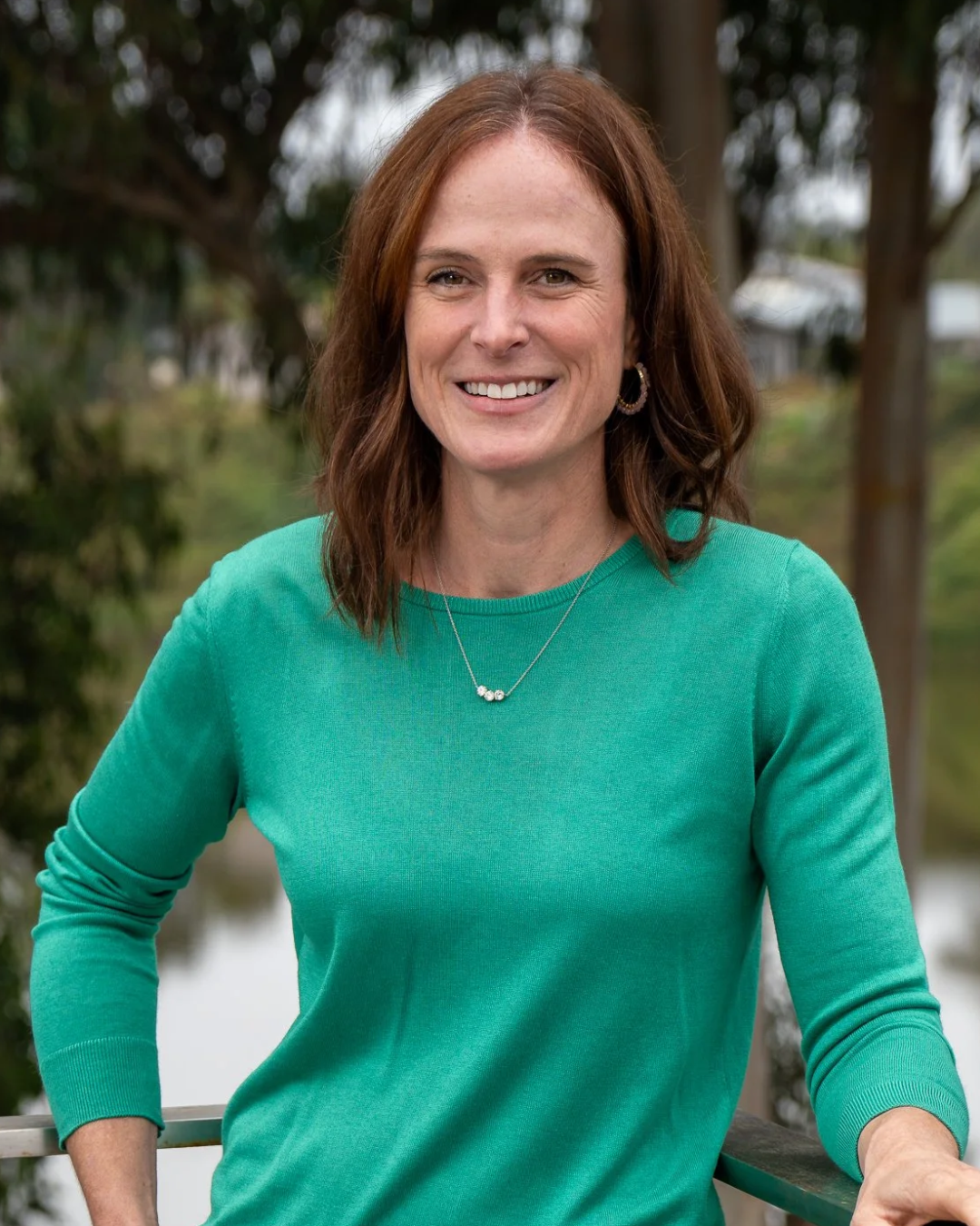 A woman with shoulder-length brown hair, wearing a teal long-sleeve shirt, standing outdoors in front of trees and water, smiling at the camera.