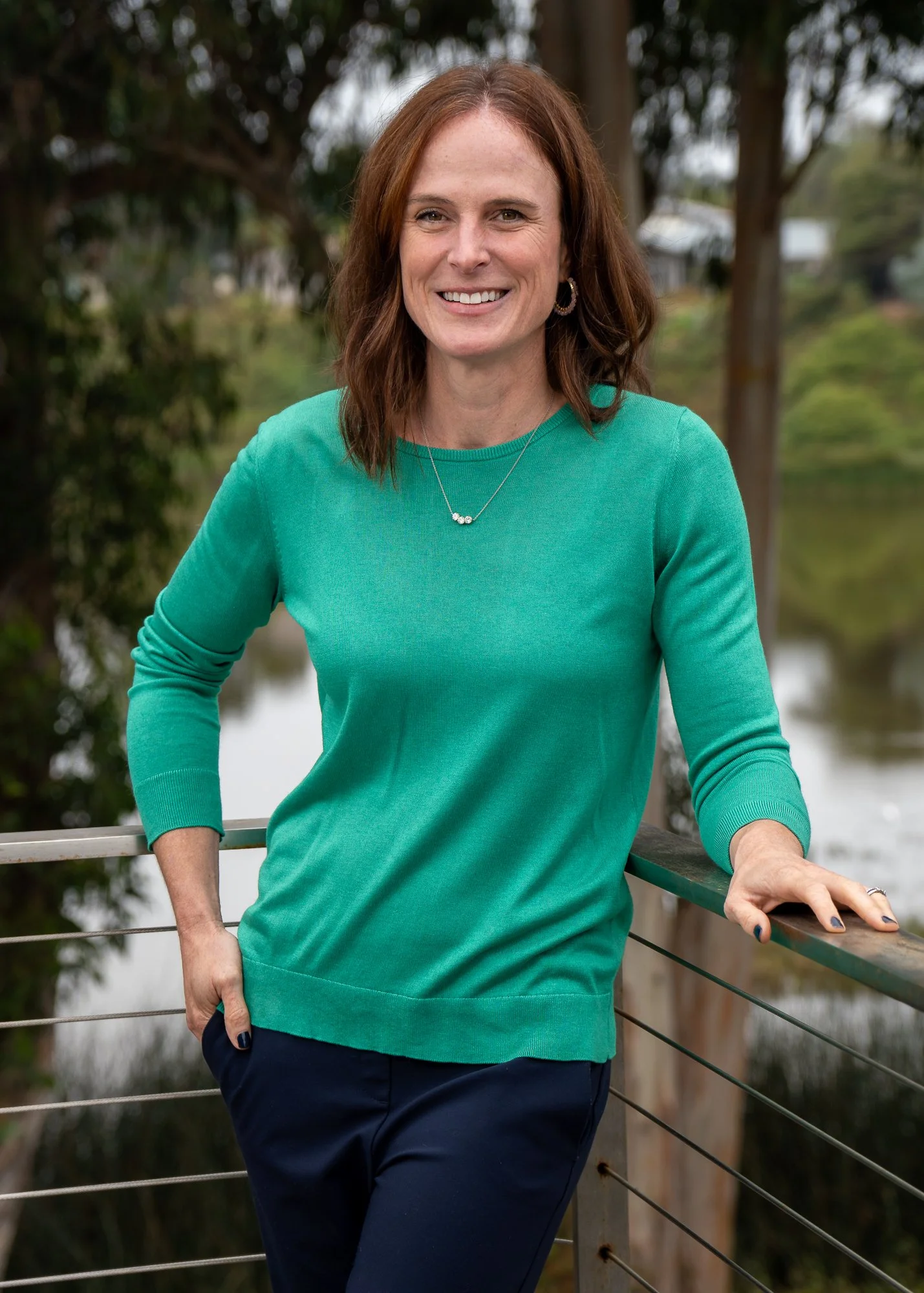 A woman with shoulder-length brown hair, wearing a teal sweater and navy pants, standing outdoors on a wooden bridge with a river and trees in the background.