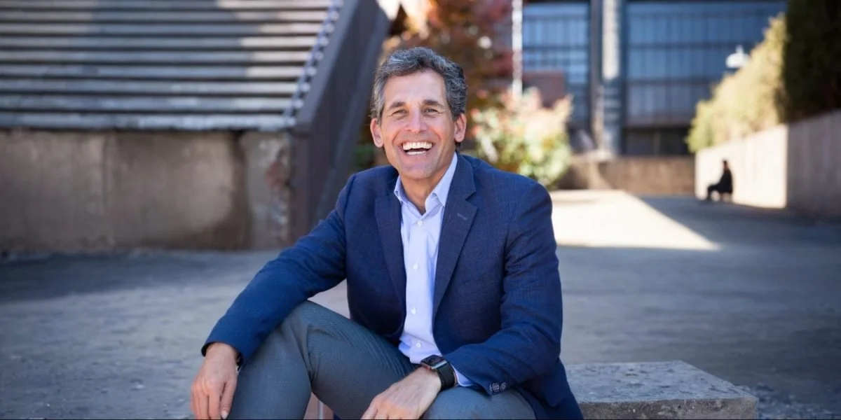 A middle-aged man in a blue blazer and light blue shirt sitting outdoors on a concrete ledge, smiling at the camera, with urban buildings and trees in the background.