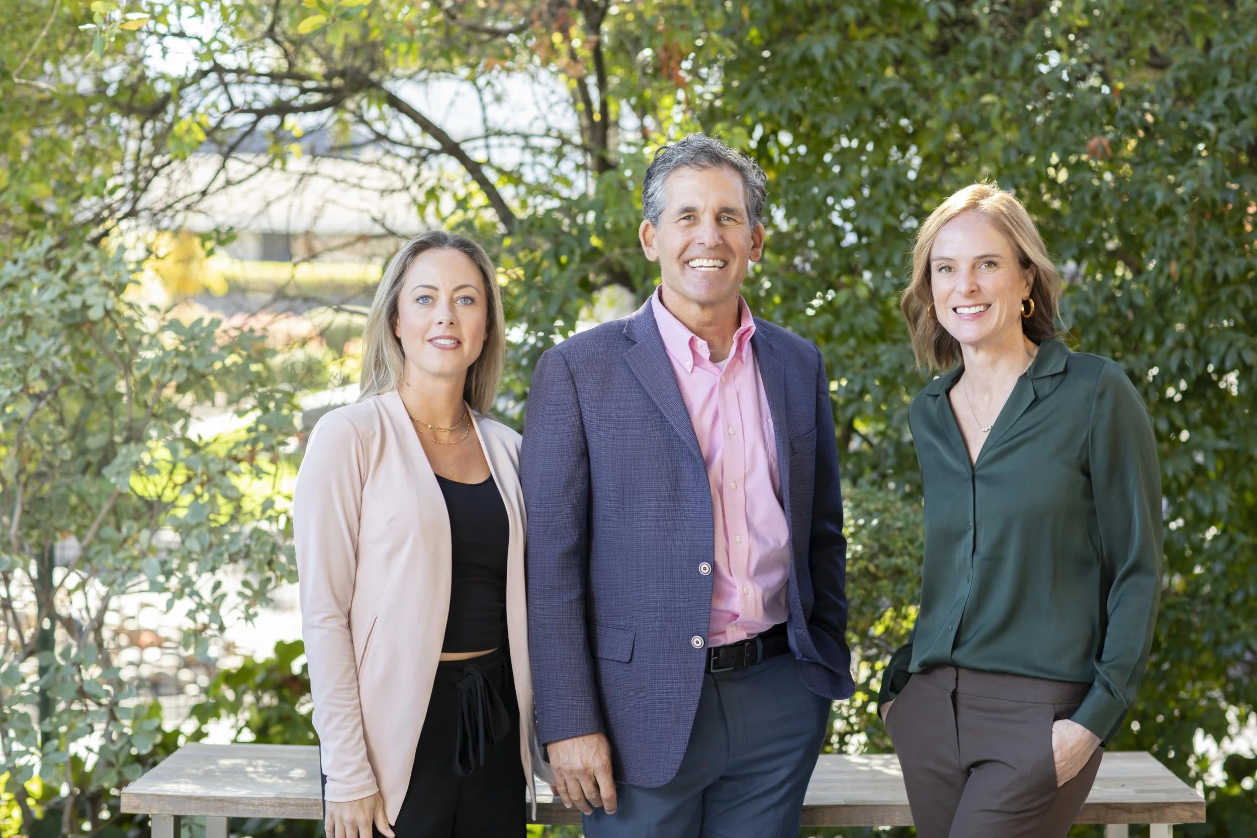 Four professionally dressed adults standing outside in front of green foliage, smiling at the camera.
