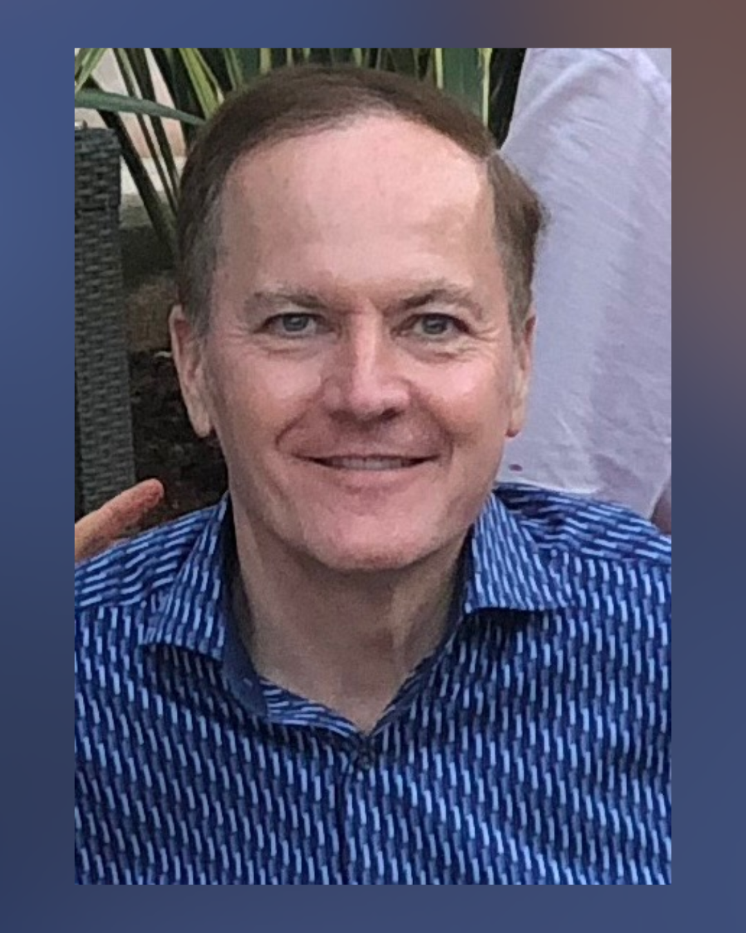 A smiling man with short light brown hair wearing a blue patterned shirt, posing outdoors in front of greenery and wicker furniture.