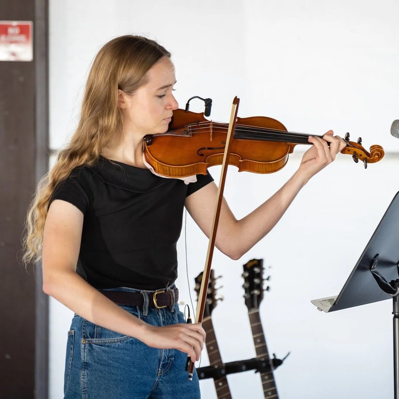 Young woman with long, wavy blonde hair playing the violin during a music practice session, standing in a room with several guitars in the background.