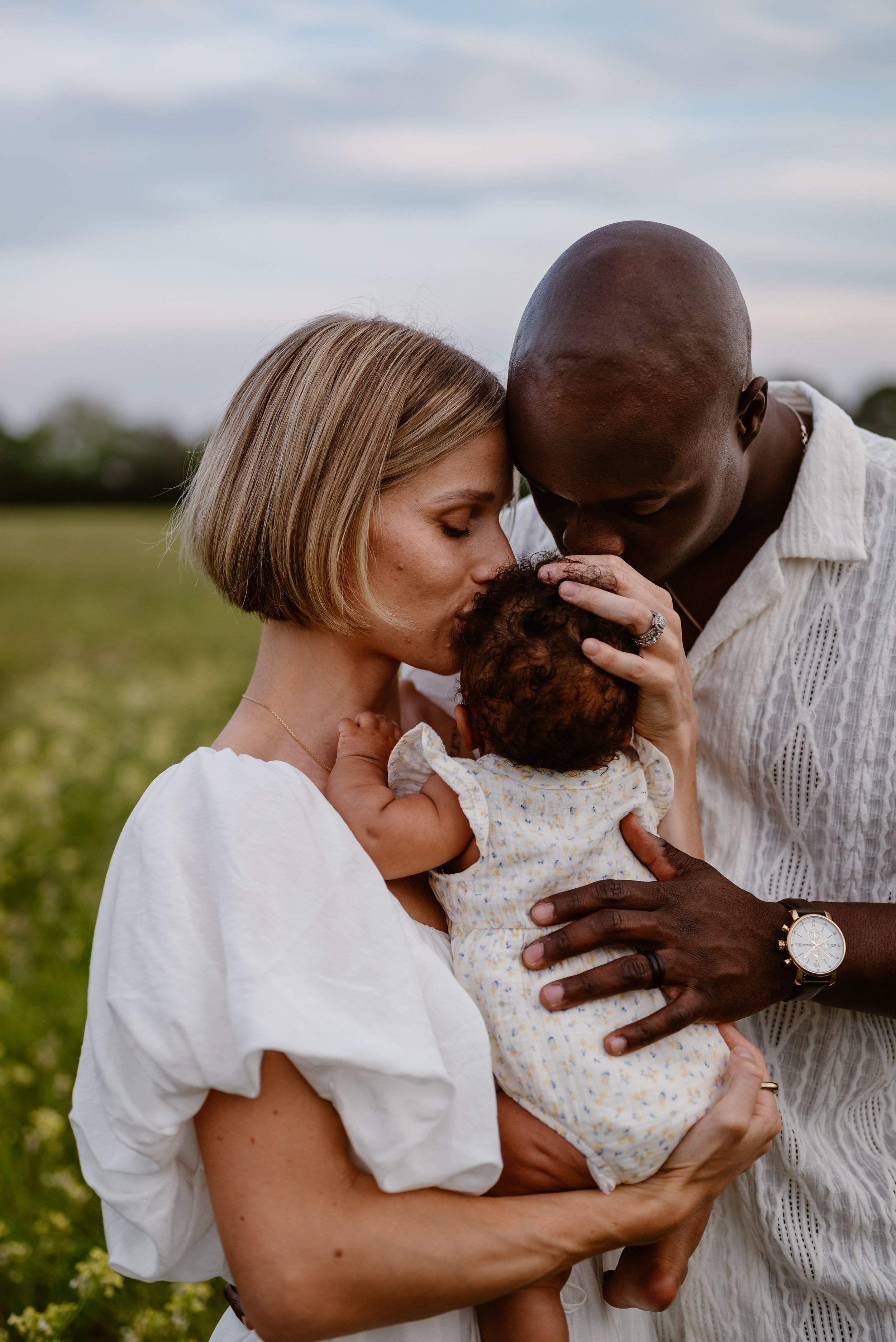 A diverse family, including a woman, a man, and a baby, sharing a tender moment outdoors with a cloudy sky and green field in the background.