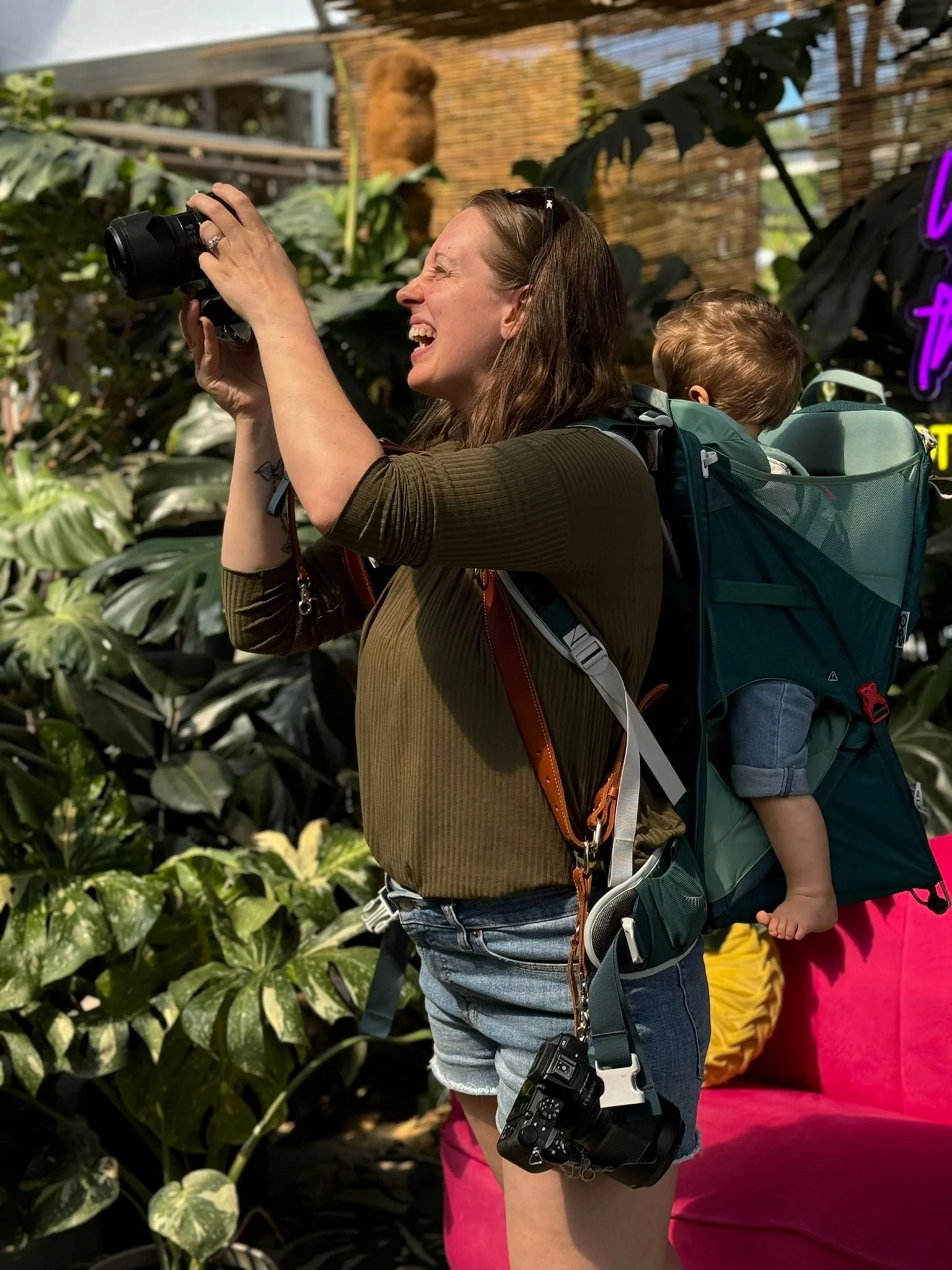 Megan Steele Giakas takes photos while holding a baby in a backpack on her back.
