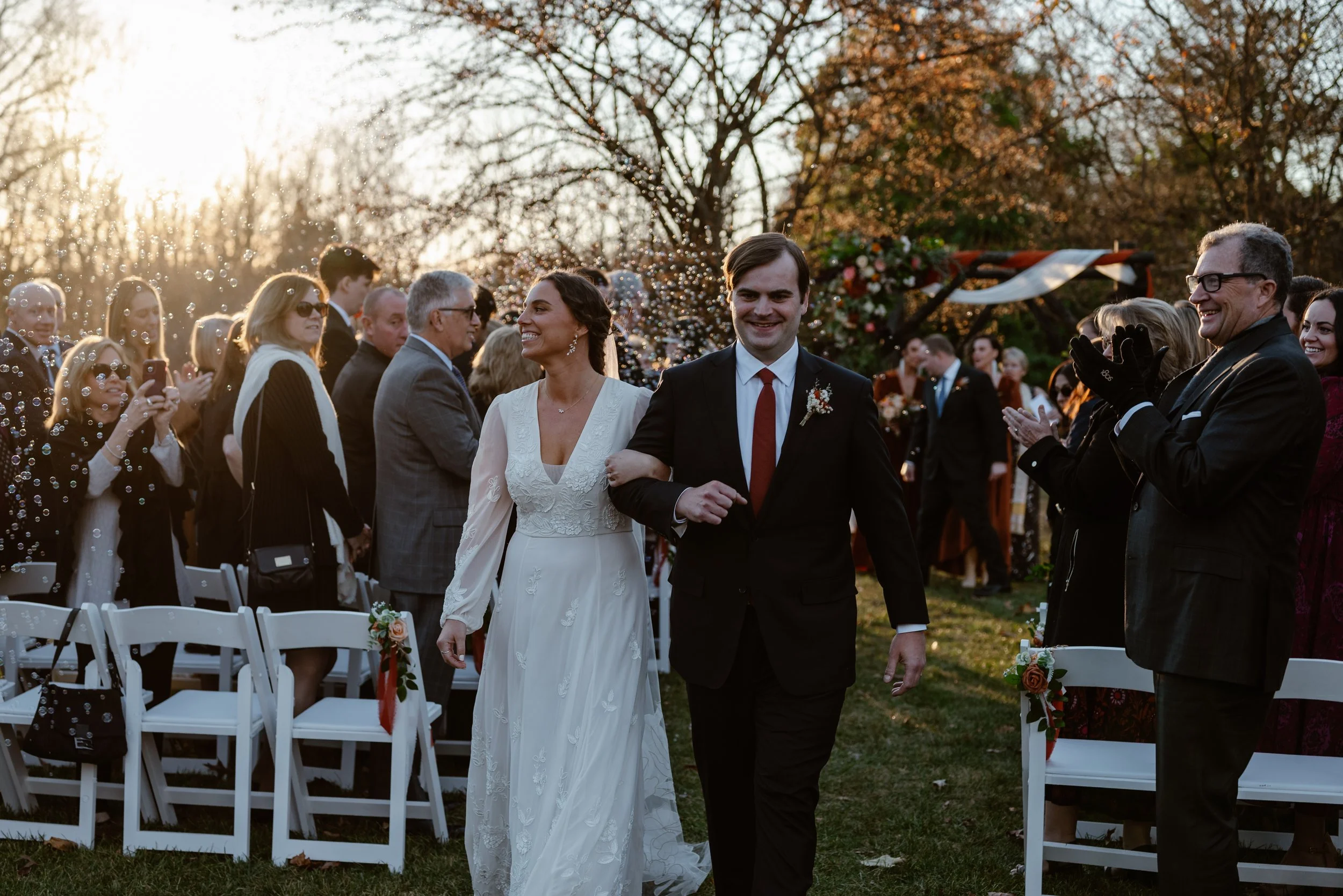 A bride and groom grin broadly while exiting their outdoor rustic fall wedding with bubbles in the air