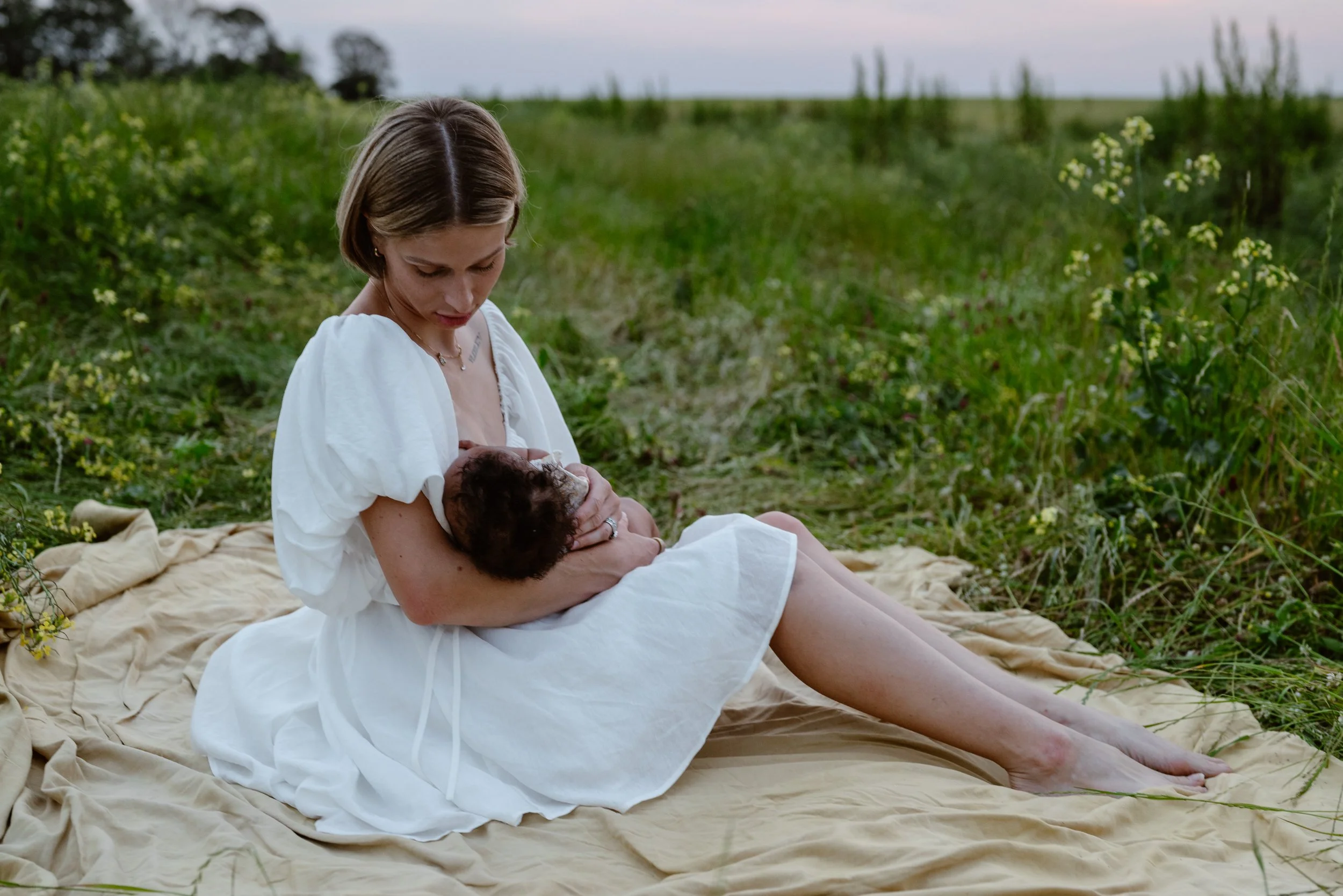 A woman in a white boho dress sitting on a blanket outdoors in a grassy field, breastfeeding a baby.