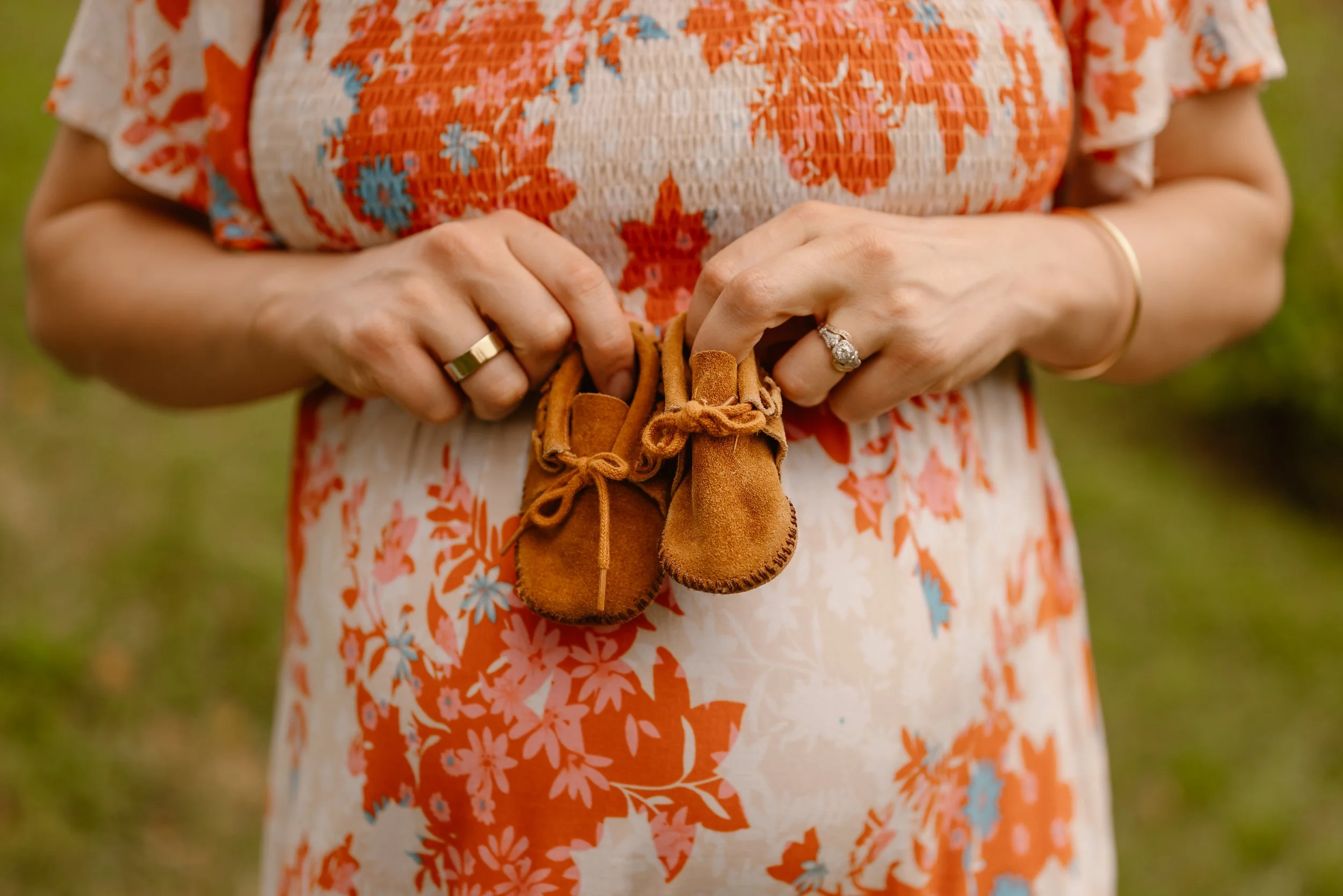 A mother holding tiny baby shoes in front of her pregnant belly, wearing a floral dress and rings during an outdoor maternity photoshoot.