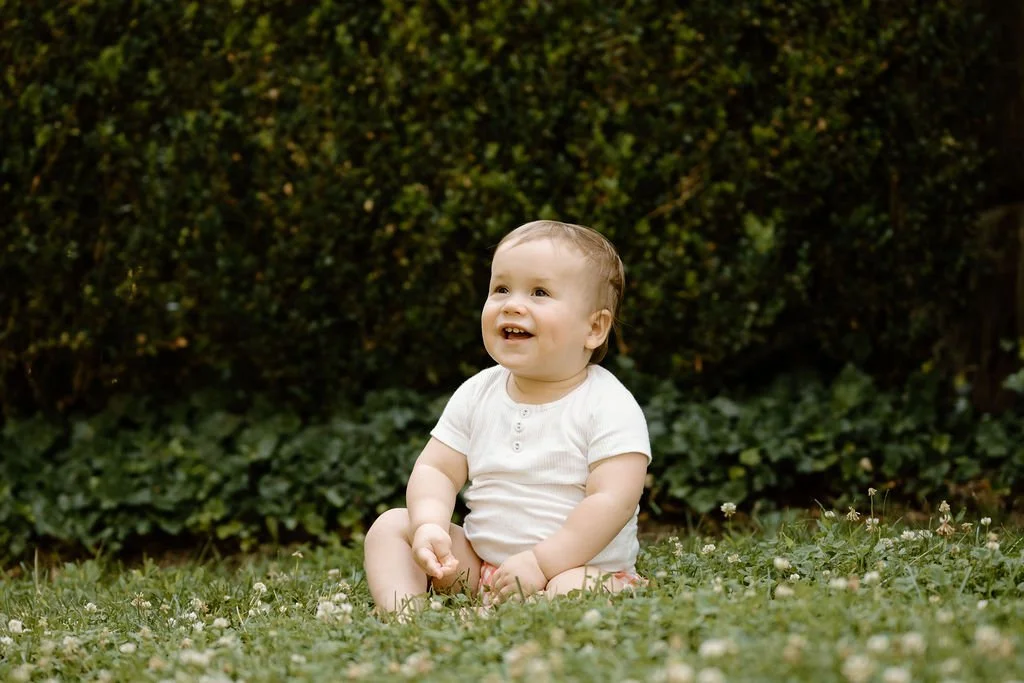 A smiling toddler sitting on grass in front of a leafy hedge, wearing a white shirt and shorts.