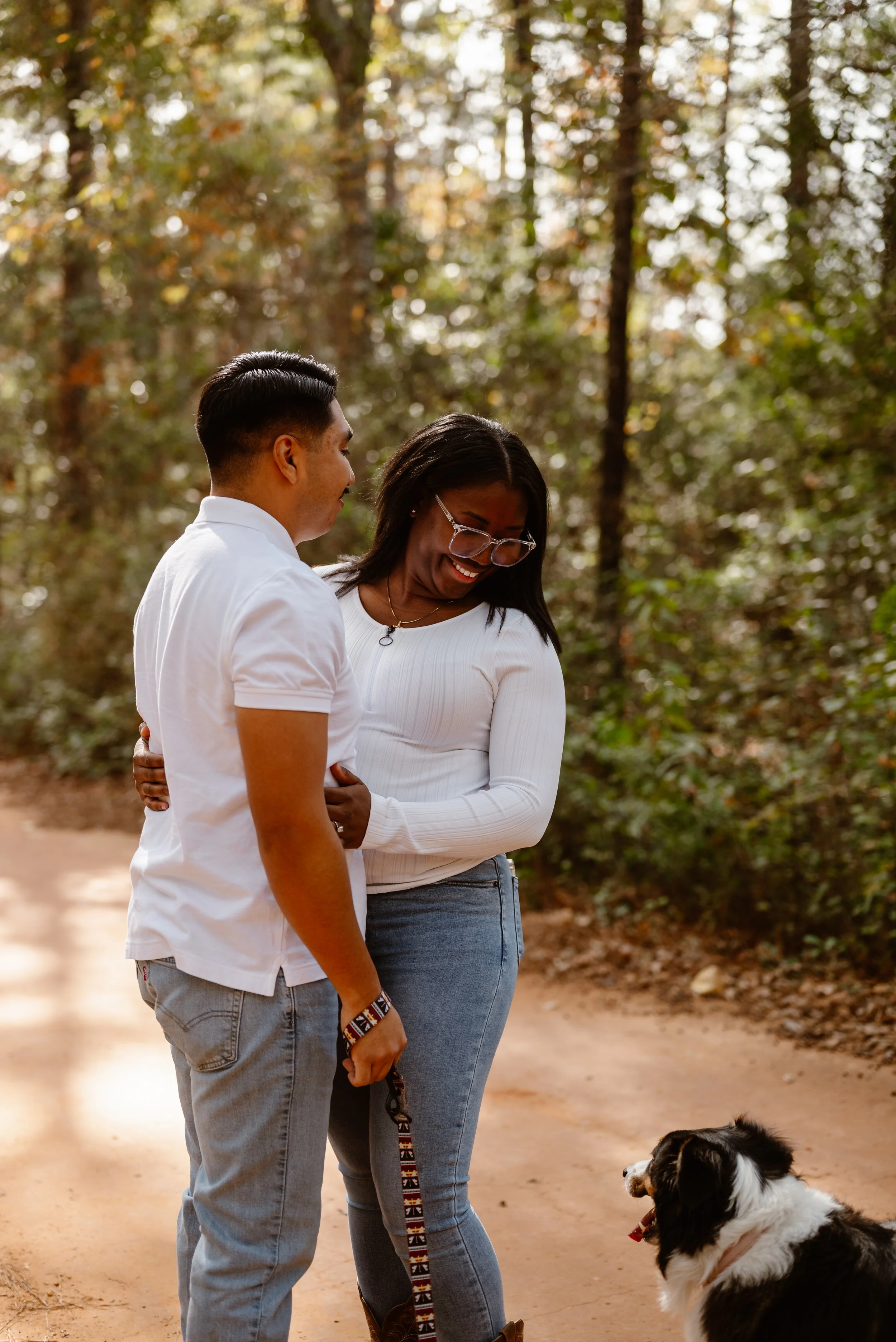 A diverse couple smiles at their dog during an outdoor engagement photoshoot