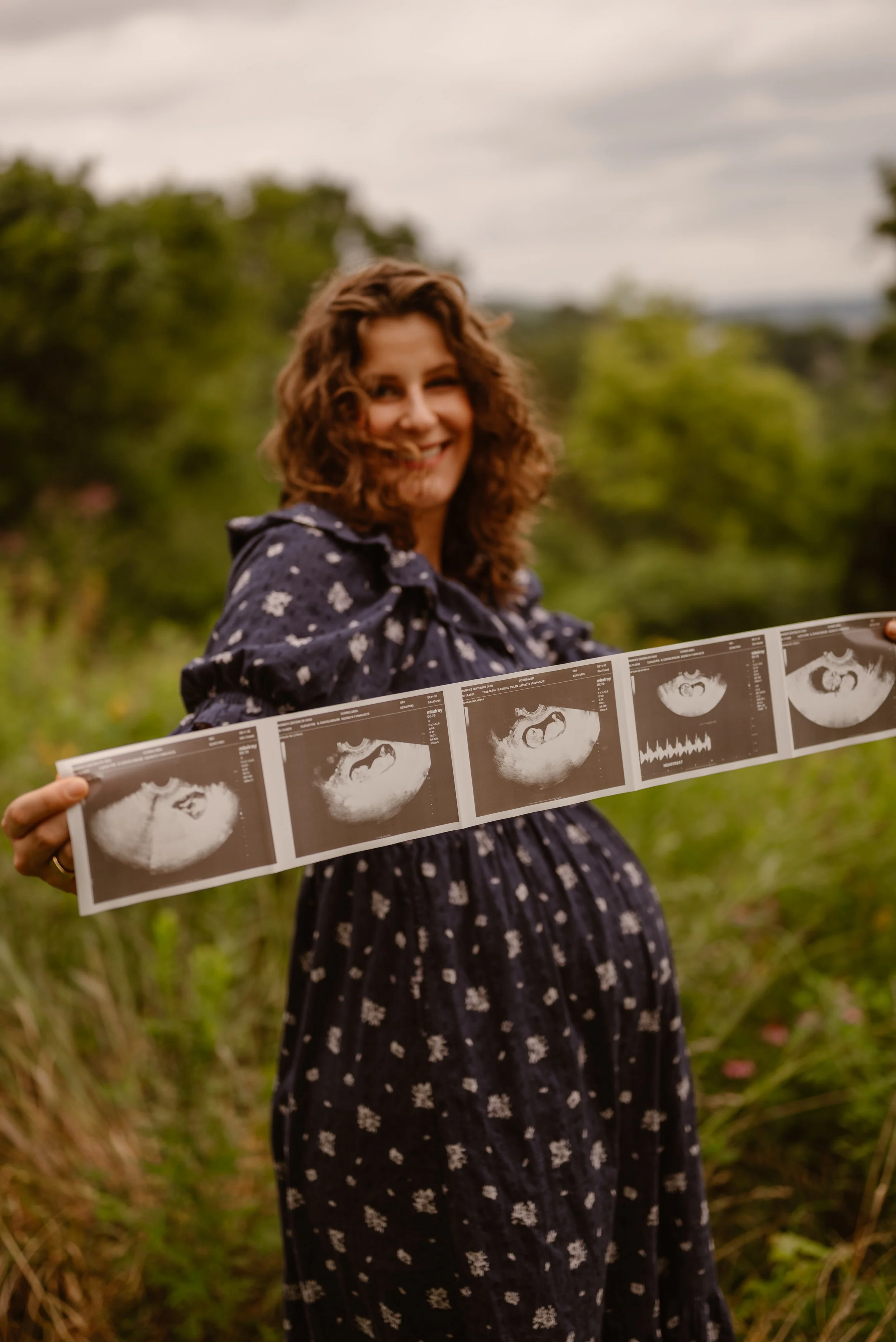 A woman holding ultrasound photos outdoors in a green field during a rustic outdoor maternity photoshoot.