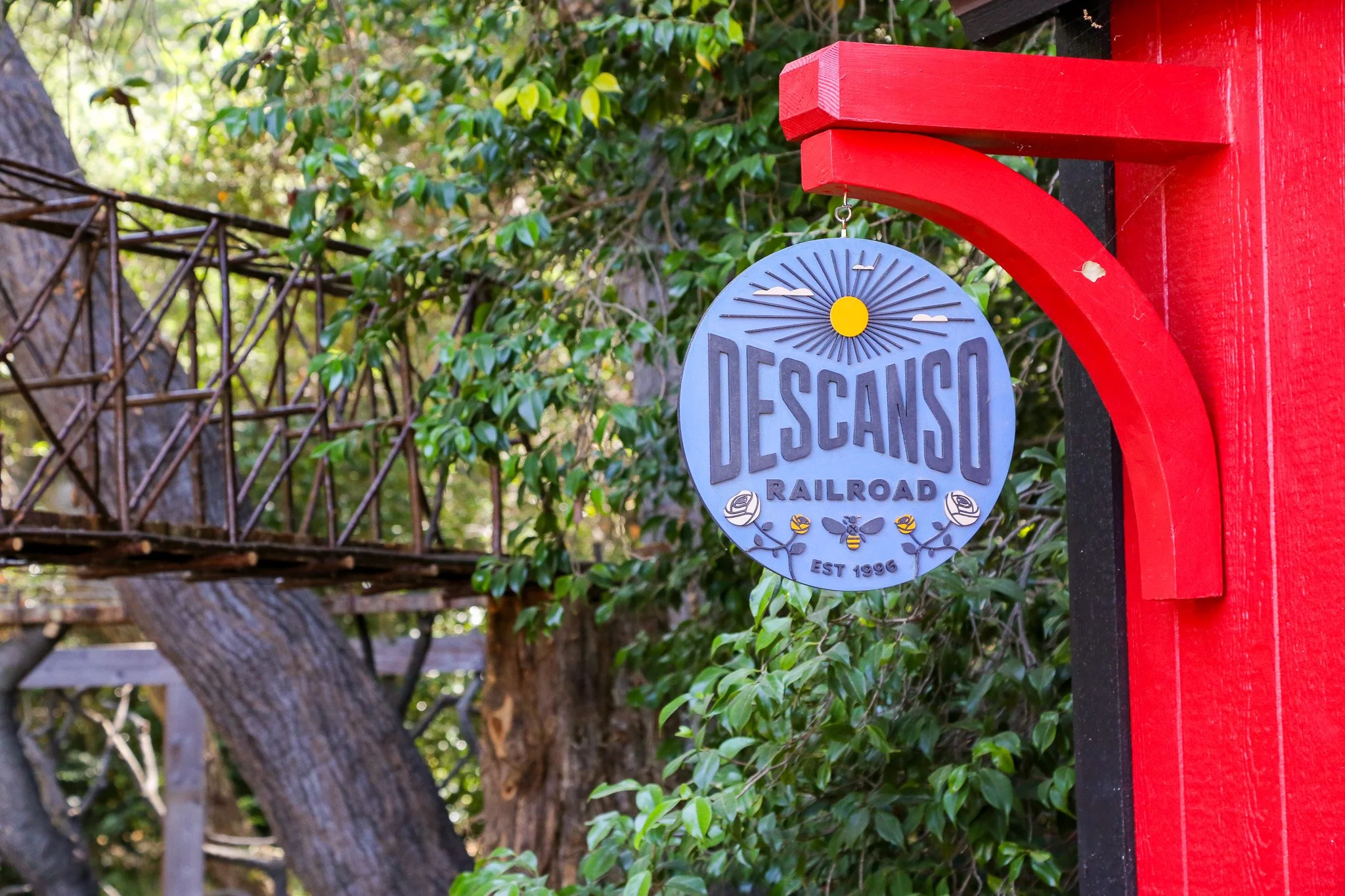 A circular sign hanging from a red post that reads 'Descanso Railroad, Est 1996' with a sun and bee design, in front of green bushes and a wooden bridge.