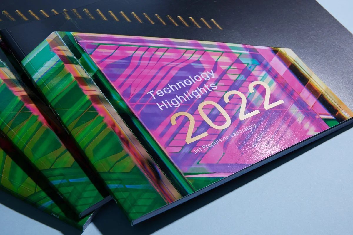 A report titled 'Technology Highlights 2022' by Jet Propulsion Laboratory, with colorful, futuristic cover art featuring geometric patterns and neon colors, is shown on a workspace desk.
