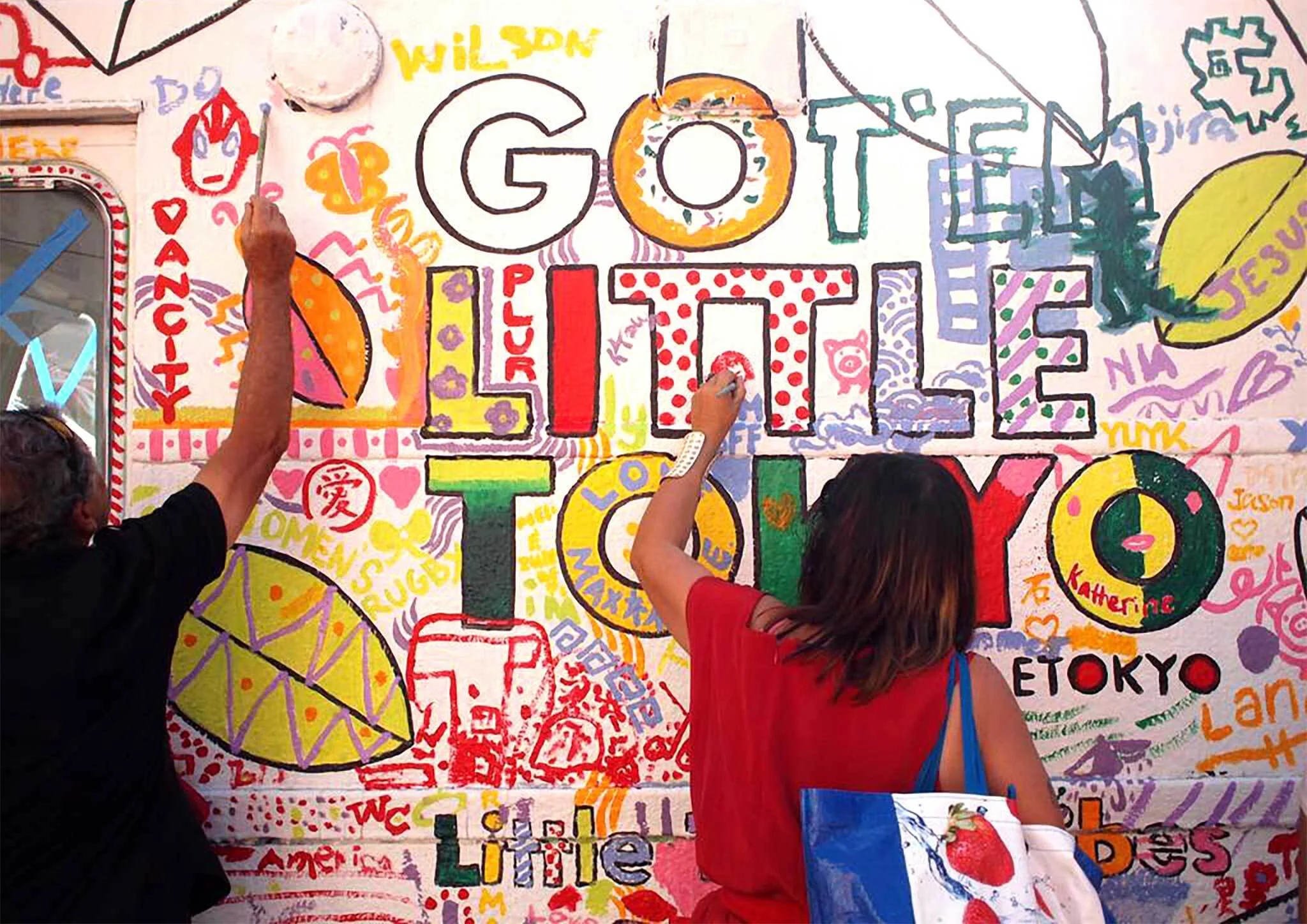 People drawing and writing on a colorful wall with the words 'GOT LIFE' in large letters at center, surrounded by various symbols, names, and illustrations.