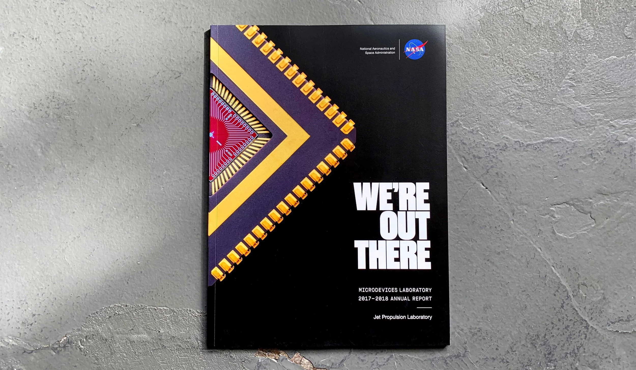 Cover page of a report titled "WE'RE OUT THERE" from NASA, related to Microdevices Laboratory, for the 2017-2018 year, with a colorful geometric design on a black background.