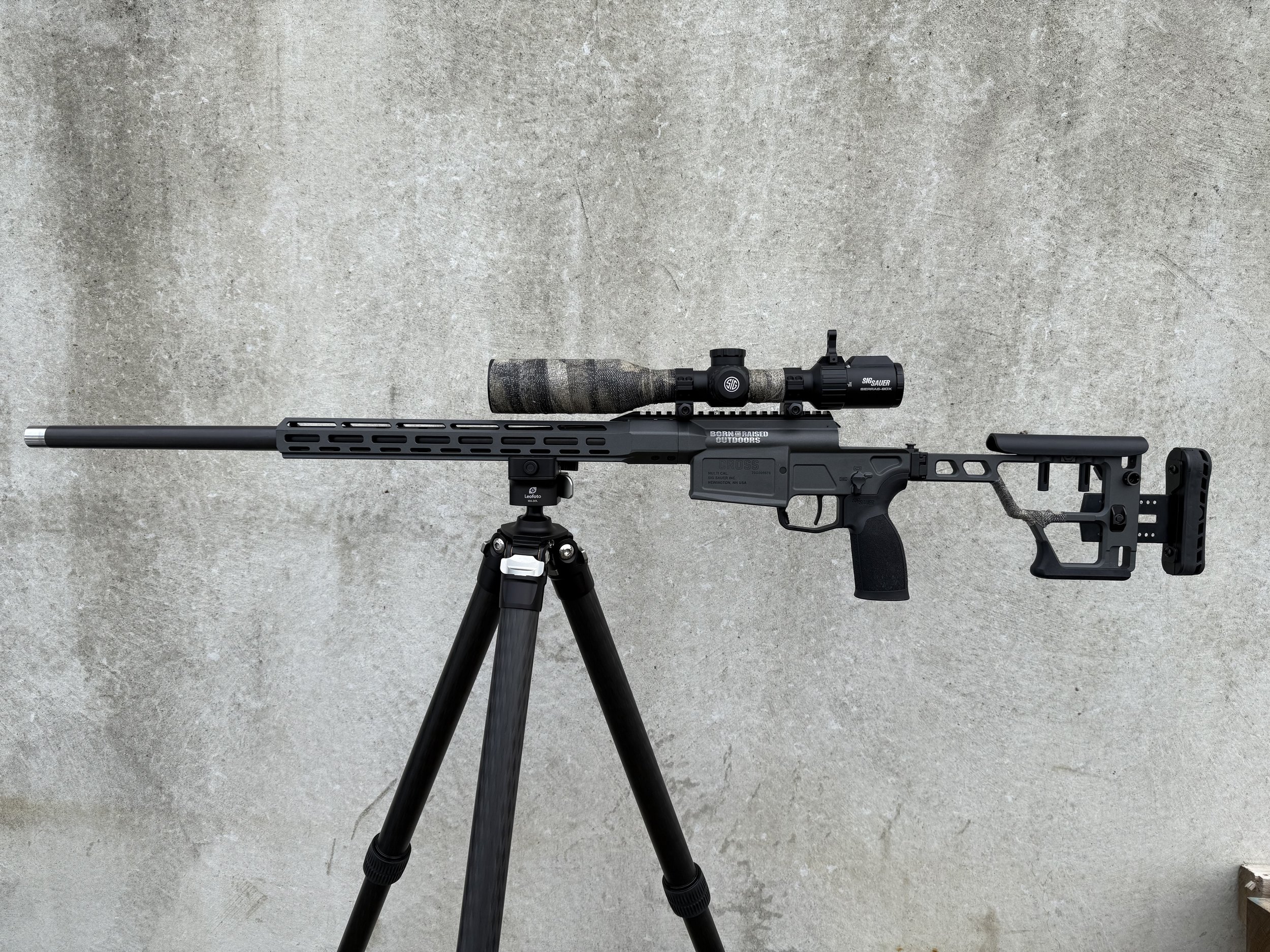 A black precision rifle with a large telescopic scope mounted on a tripod, set on a concrete surface.