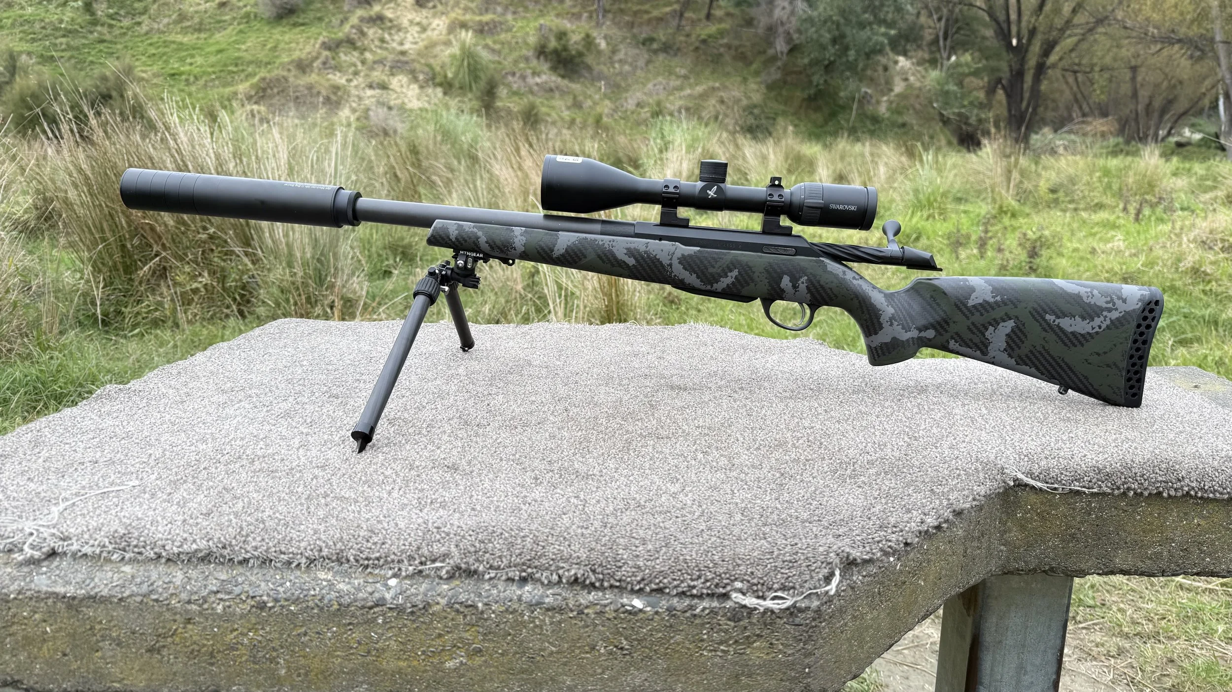 A rifle with a camouflage pattern and a scope mounted on top, placed on a bipod stand on a concrete surface outdoors in a grassy and wooded area.