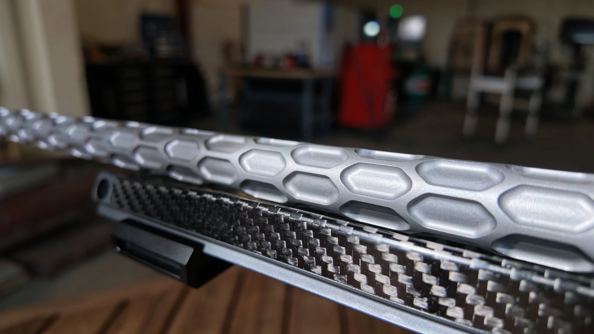 Close-up of a metallic object with a honeycomb pattern and a section of carbon fiber material, with a blurred background of a room with furniture and various items.