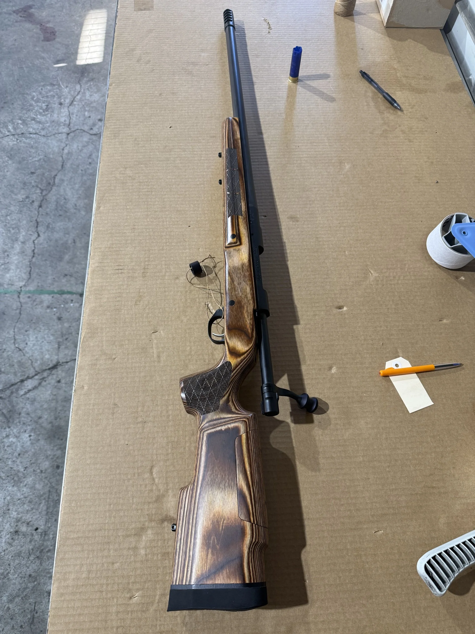 A rifle with a wooden stock and black barrel lying on a brown cardboard surface, surrounded by a few pens, a blue shotgun shell, a small roll of tape, and a white vent on the bottom right corner.