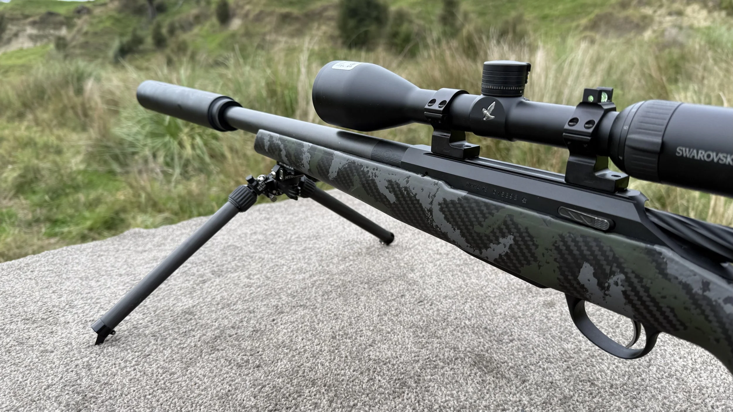 A sniper rifle with a scope mounted on a tripod, resting on a concrete surface outdoors with a grassy landscape in the background.