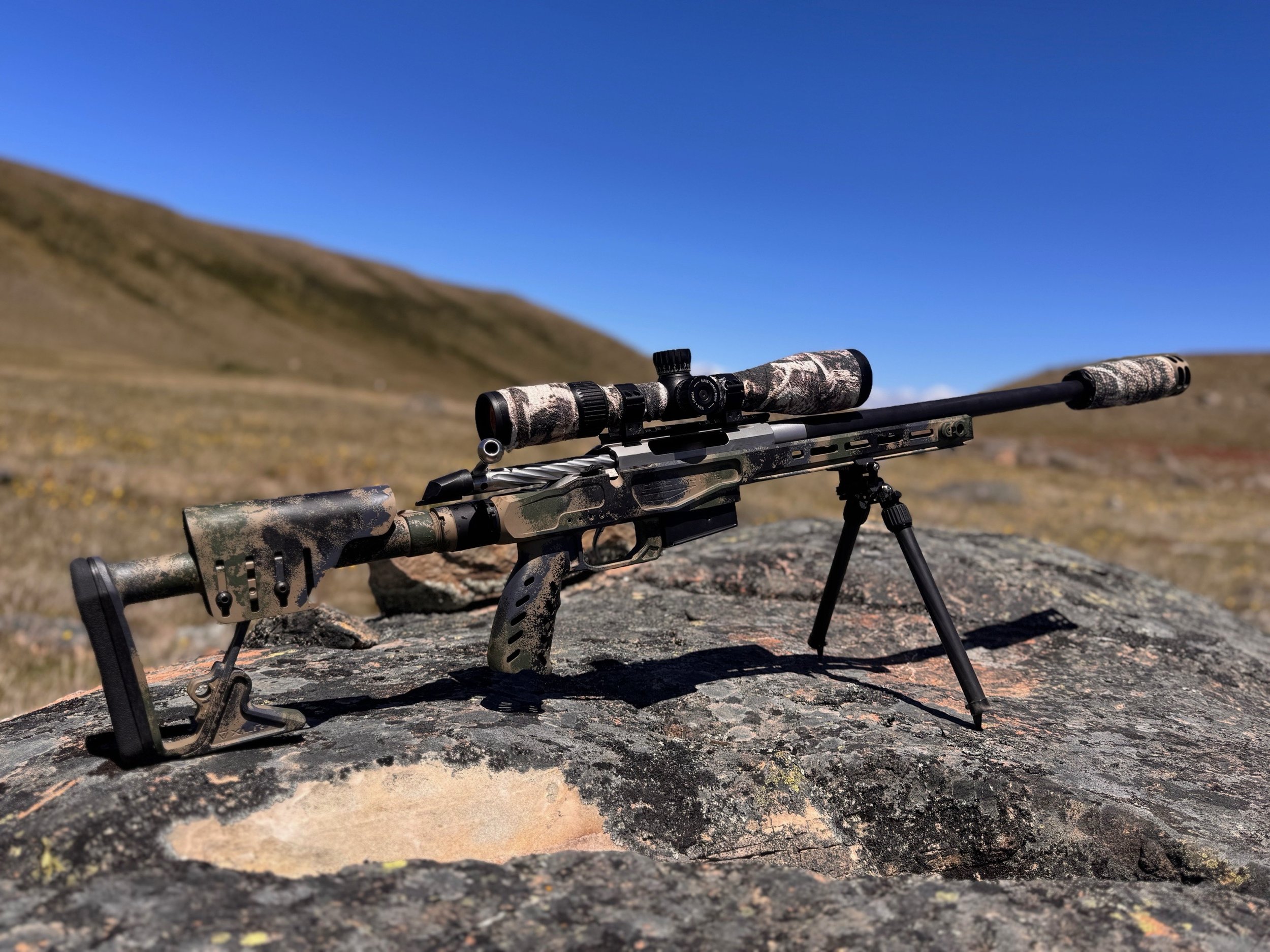 A camouflage-patterned sniper rifle with a scope and bipod resting on a rocky surface in an open landscape with rolling hills under a clear blue sky.